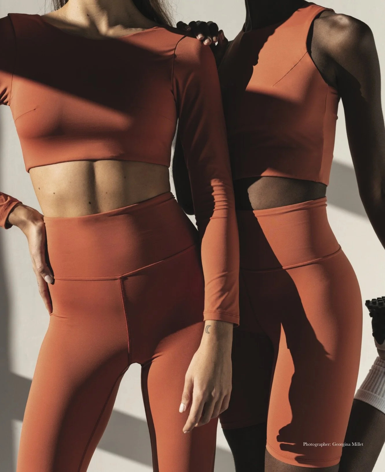 Two women wearing matching burnt orange workout outfits, including leggings and crop tops, standing close together in a minimalistic setting with shadows cast on their bodies.