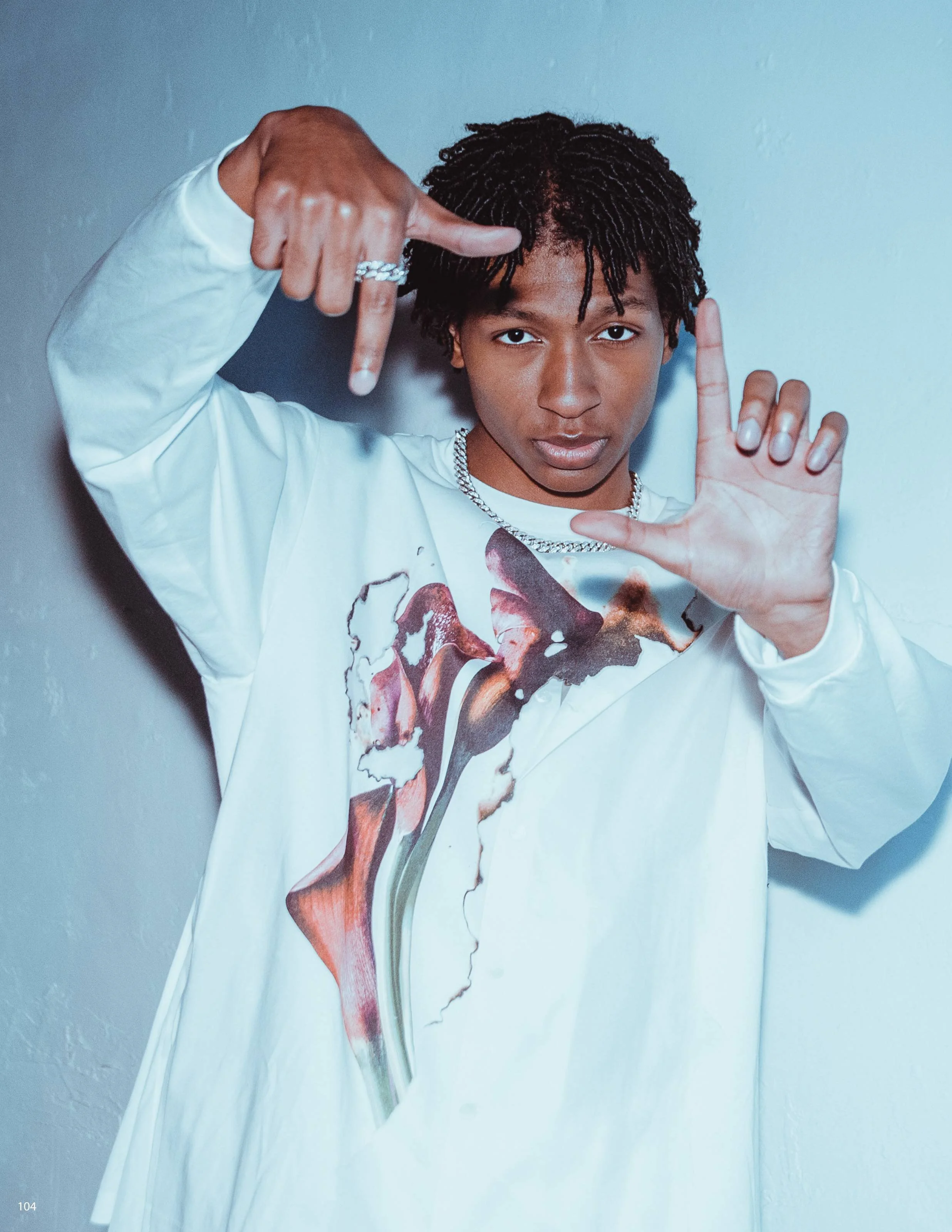 Young person with dreadlocks wearing a white long-sleeve shirt, pointing with both hands and making a framing gesture, standing against a plain light-colored wall.