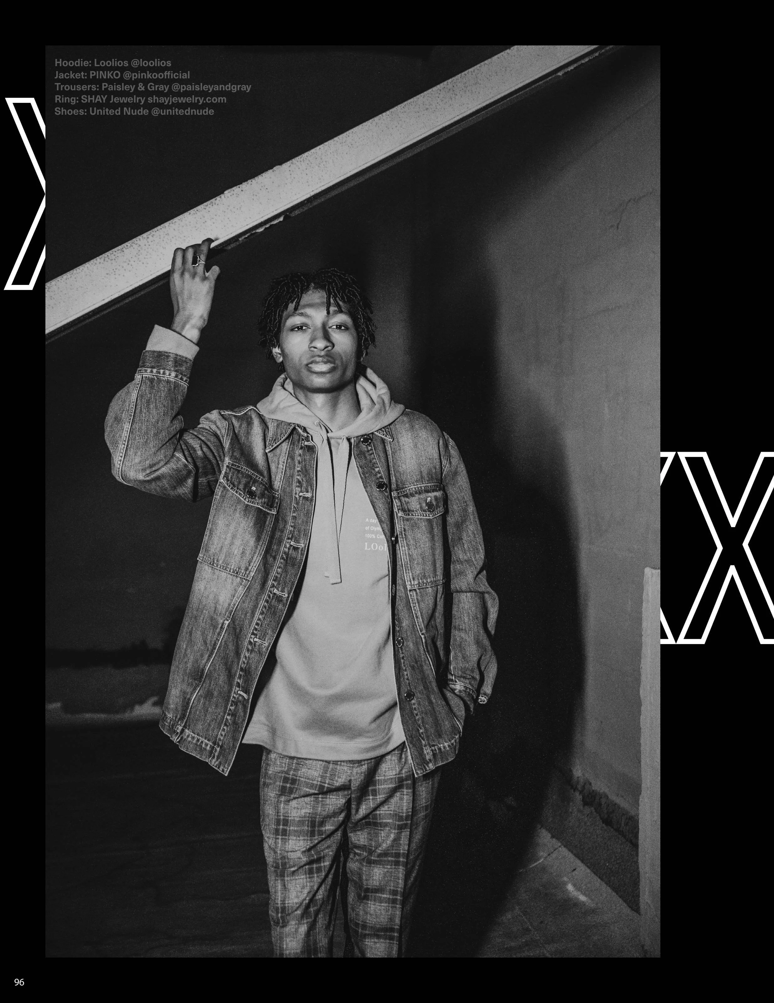 Black and white photo of a young man standing outdoors at night, wearing a denim jacket over a hoodie and plaid pants, with one hand raised to touch a ledge above him.