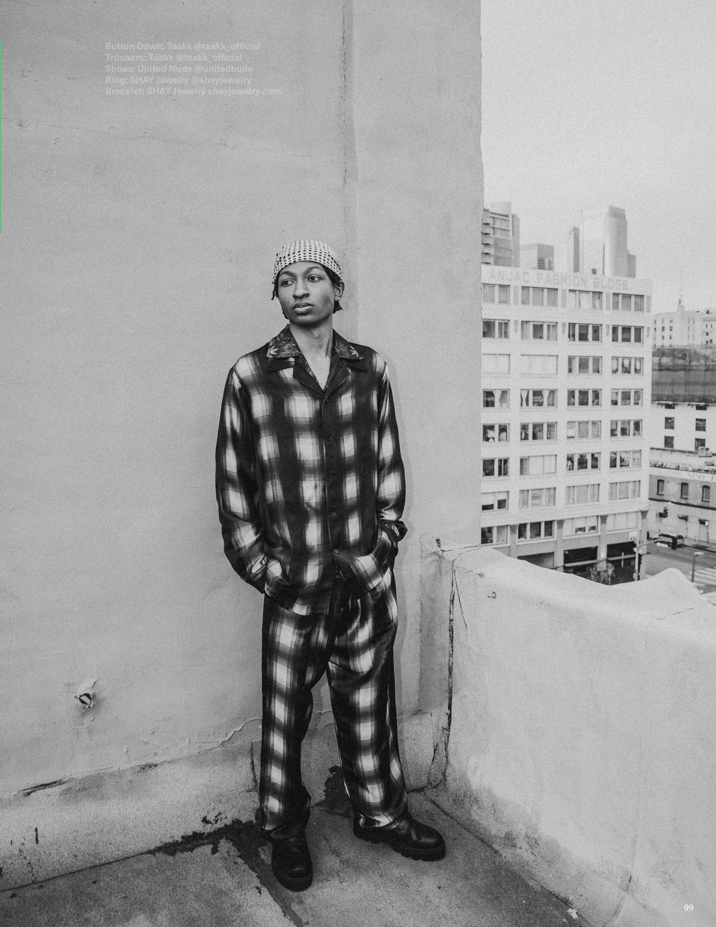 A young person standing against a concrete wall on a rooftop, wearing a plaid shirt, matching plaid pants, black boots, a patterned cap, with city buildings in the background.