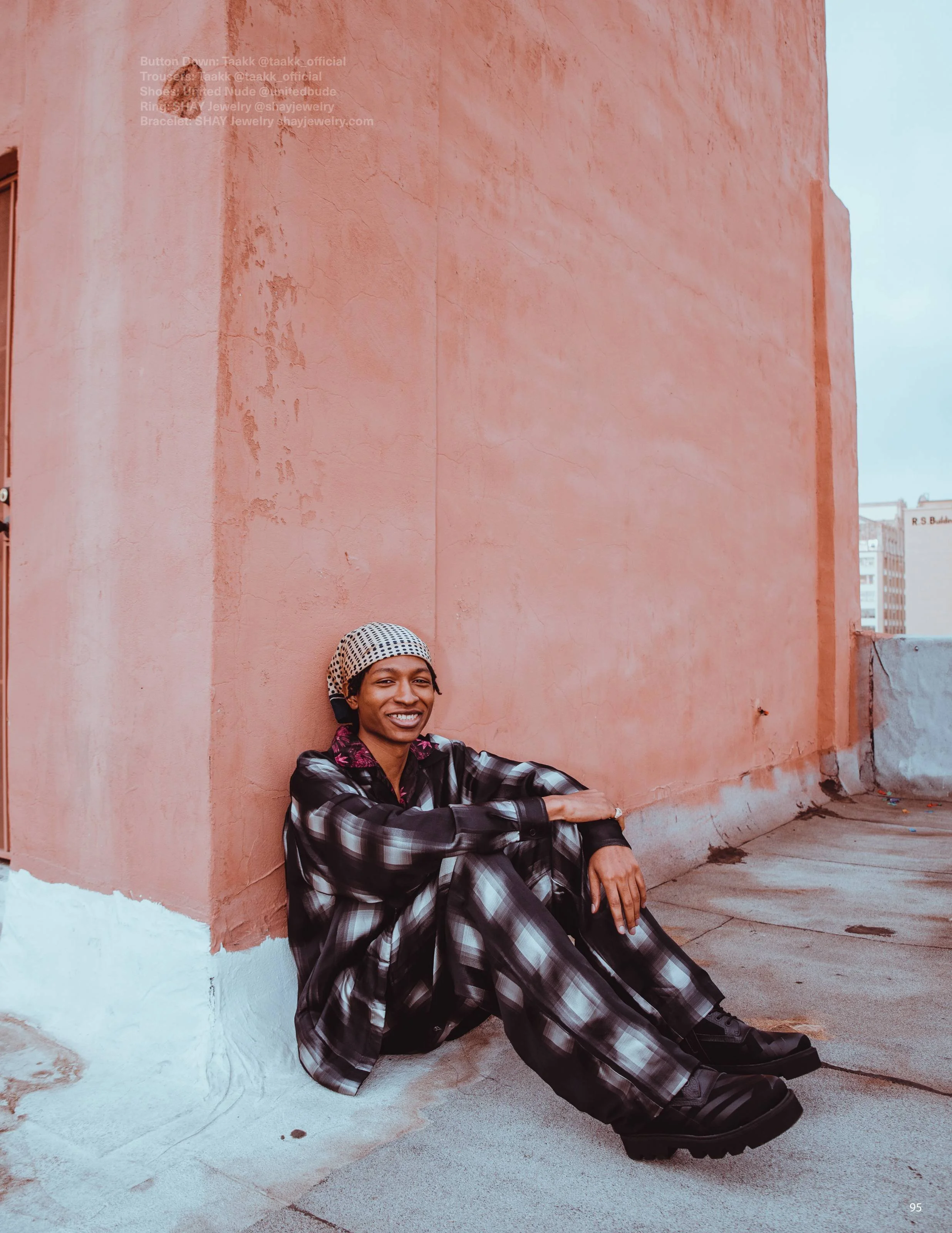 A person sitting on the ground next to a pink textured wall, smiling at the camera, wearing a checkered dress, black combat boots, and a headscarf.