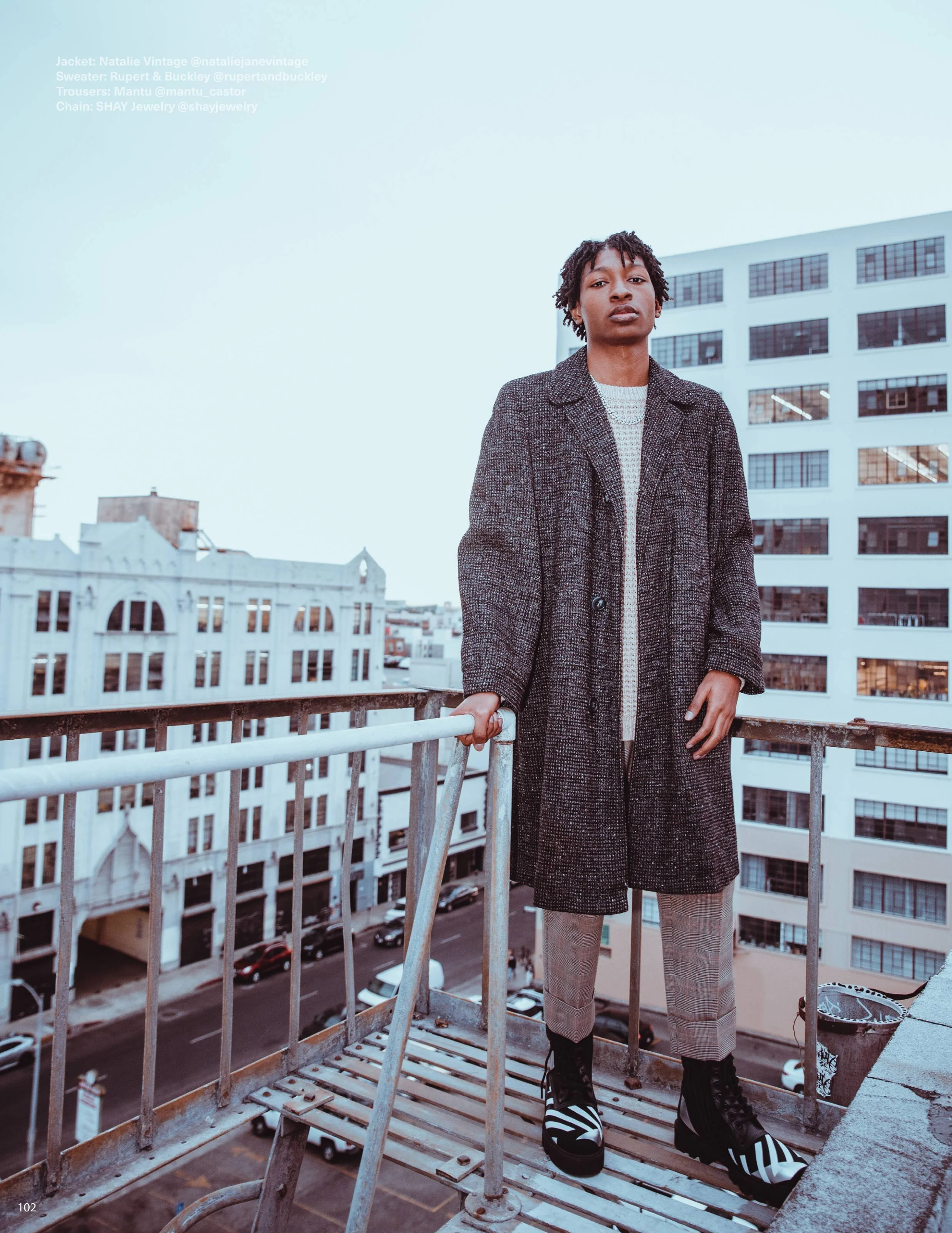 A person standing on a rooftop balcony with a tall building in the background, wearing a long checkered coat, grey pants, and black and white striped platform shoes.