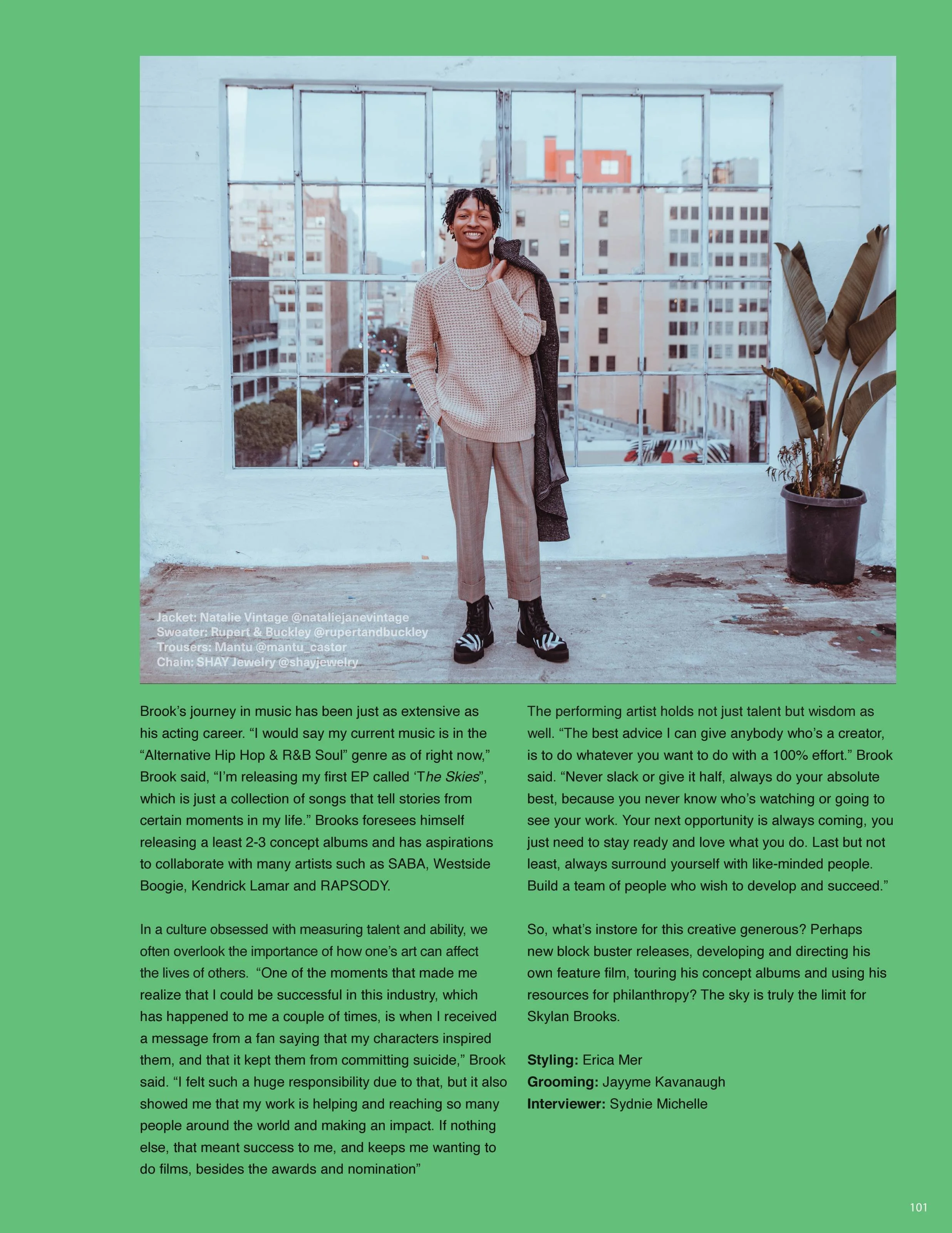 Image of a person standing in front of a large window with a cityscape view, smiling, dressed in trendy clothing, with a potted plant on the side, and a green border around the image.