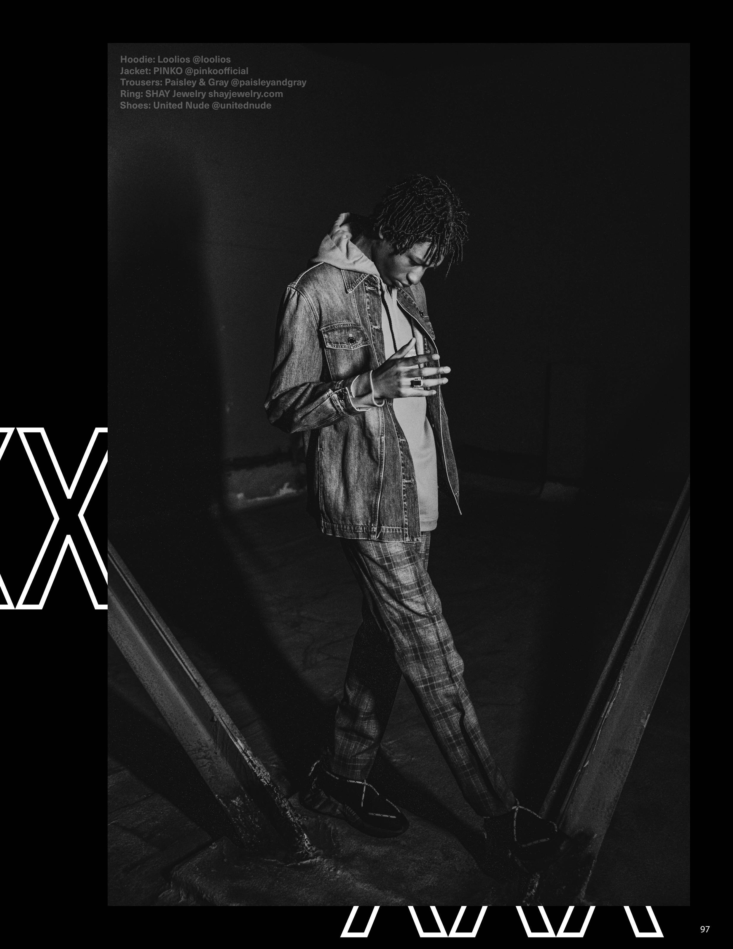 Black and white photo of a young man with dreadlocks looking down while standing inside a metallic structure. He is wearing a denim jacket over a hoodie, plaid pants, and boots.