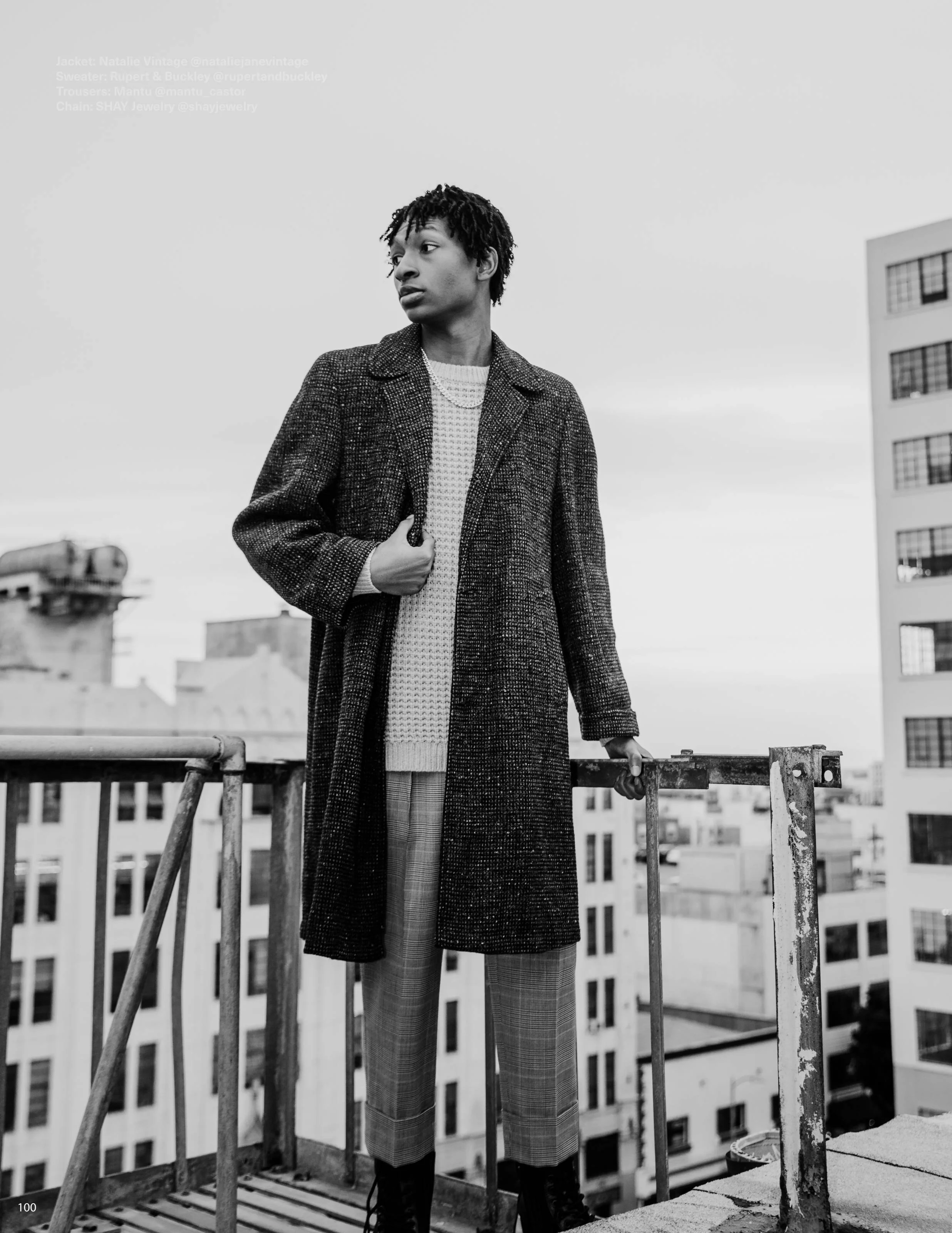 Black and white photo of a young person standing on a rooftop balcony, wearing a vintage coat, sweater, plaid pants, and chain jewelry, with buildings in the background.