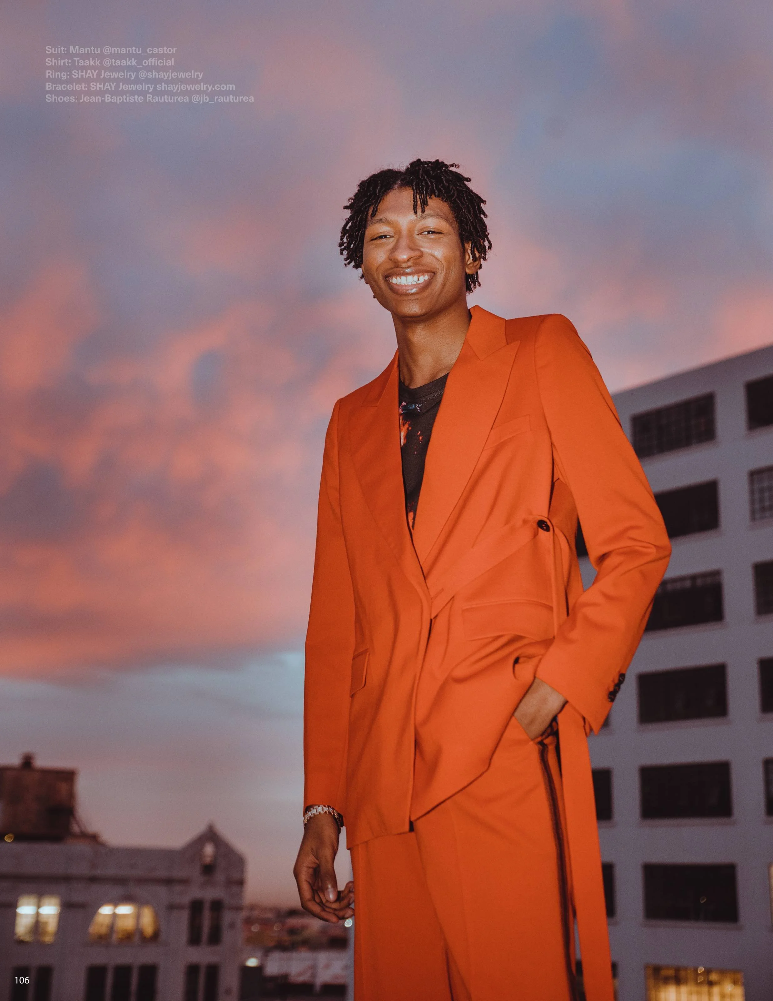 A man wearing an orange suit and black shirt stands outdoors during sunset, smiling and showing his teeth, with a background of a pink and purple sky and residential buildings.