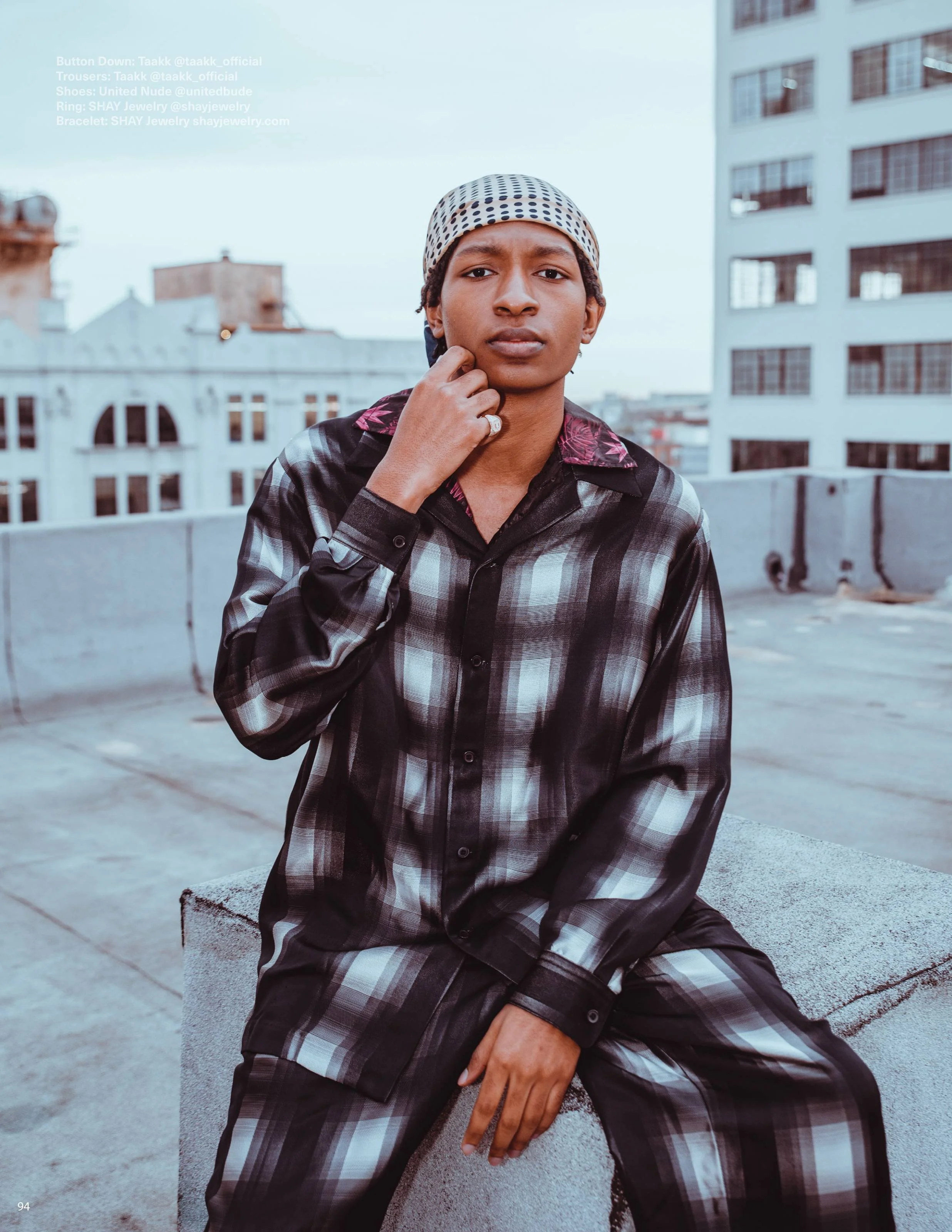 A young man with short hair wearing a checkered shirt and a polka dot cap is sitting on a city rooftop with tall buildings in the background. He has a serious expression and is touching his chin with his hand, which has a ring on the finger.