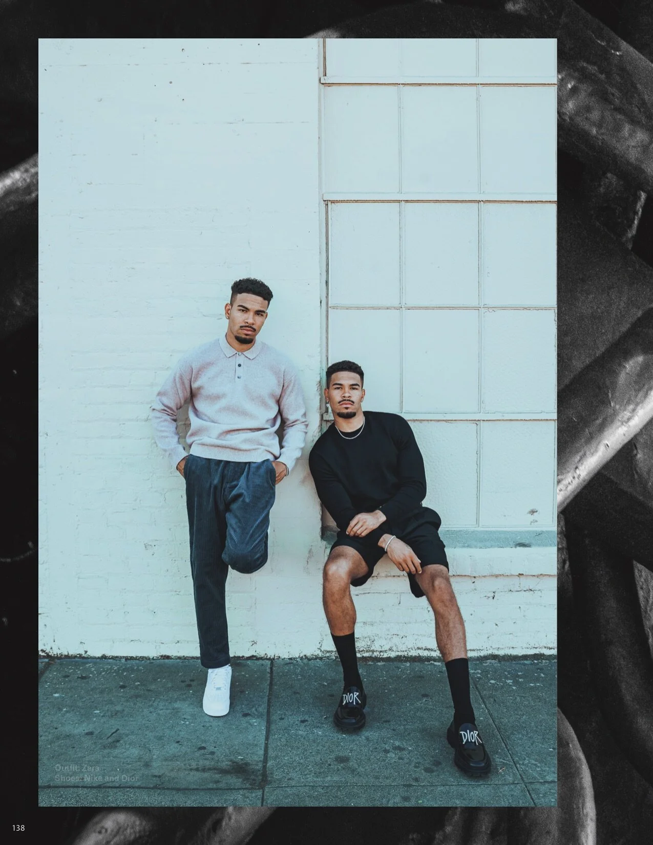 Two young men posing against a white wall, one standing with hands in pockets and one sitting with legs apart, wearing casual clothing and sneakers.