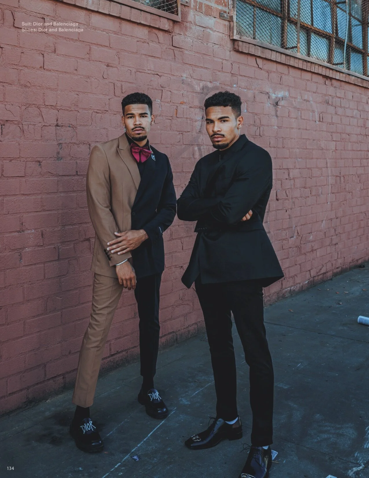 Two young men wearing suits, one in a tan suit with a pink bow tie and the other in a black suit, standing against a pink brick wall. The man in the tan suit has his hands close to his waist, and the man in the black suit has his arms crossed. Both a