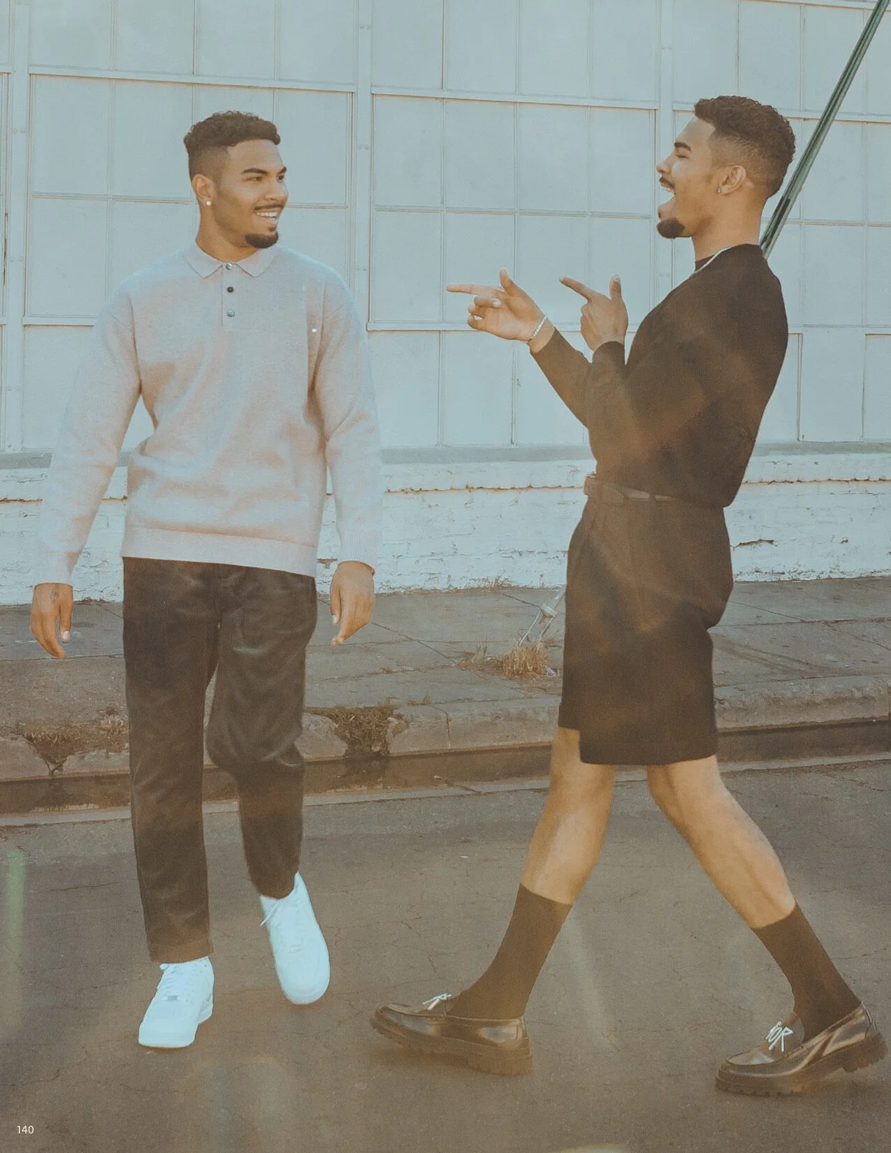 Two young men walking and talking on the sidewalk. One is dressed casually in a light sweater and dark pants, while the other wears a black jacket with a skirt and chunky shoes. They appear to be engaged in a friendly conversation.