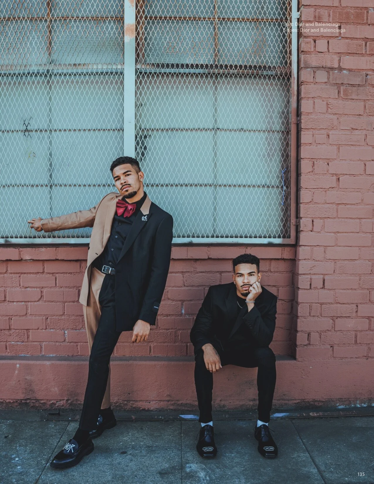Two young men in suits posing against a pink brick wall with a large window, one standing and one sitting, in an urban setting.