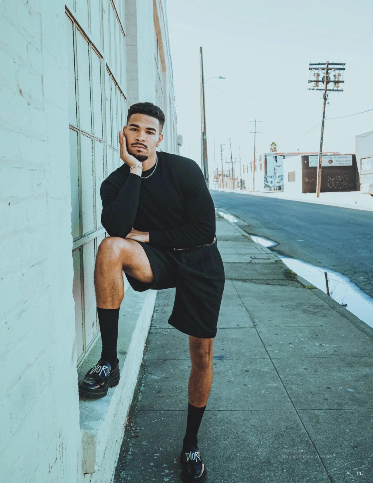 A young man with a beard and short hair poses on a city sidewalk, leaning against a pale blue brick wall, wearing all black including a long-sleeve shirt, shorts, and black Dior slides, with one leg raised on the wall and hand resting on his face, on
