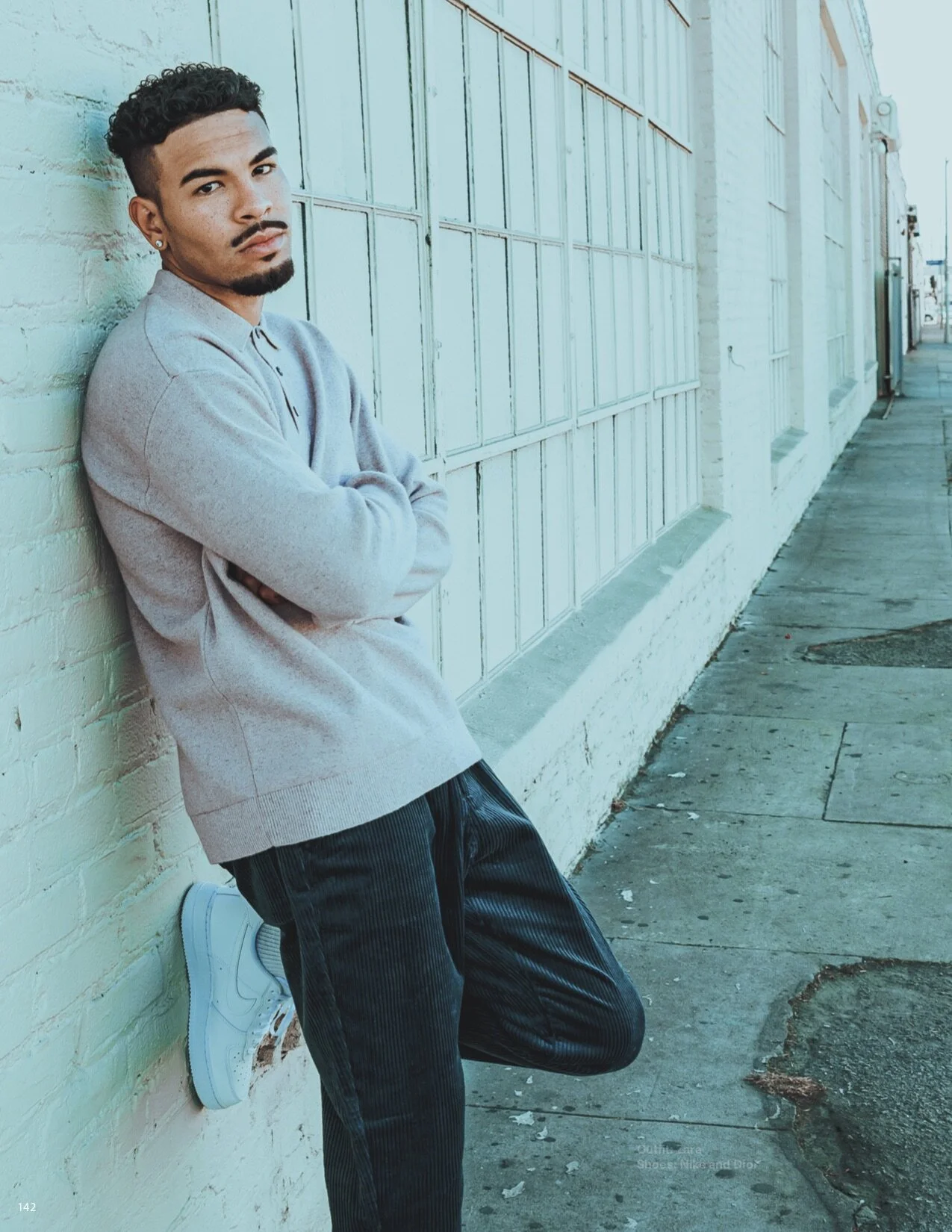 A young man with curly hair, a goatee, and wearing a beige sweater, black striped pants, and white sneakers is leaning against a white brick wall with his arms crossed, looking at the camera in an urban outdoor setting.