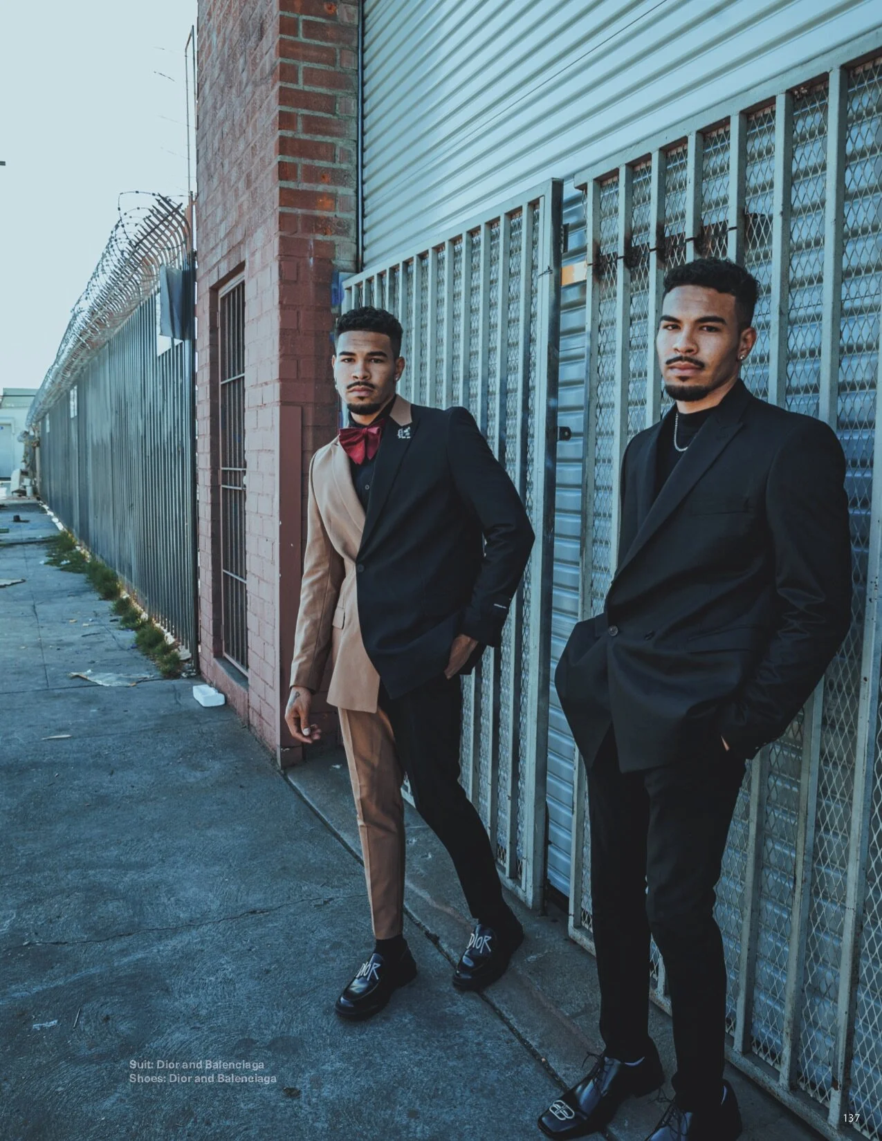 Two young men in black suits, one with a two-tone beige and black suit, the other fully black, standing outside near a brick building and metal fence on a city sidewalk during daylight.