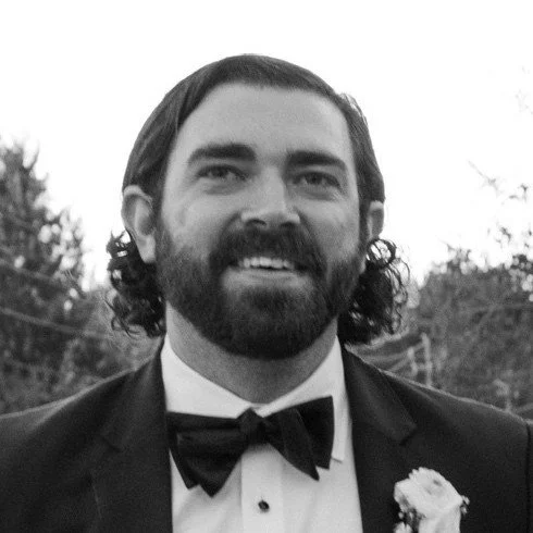 A man with a beard and curly hair wearing a tuxedo with a bowtie and a boutonniere, smiling outdoors.