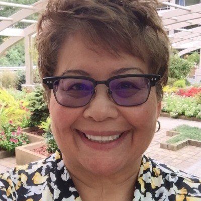 Woman wearing glasses and patterned blouse smiling in an outdoor setting with plants and a landscaped garden in the background.