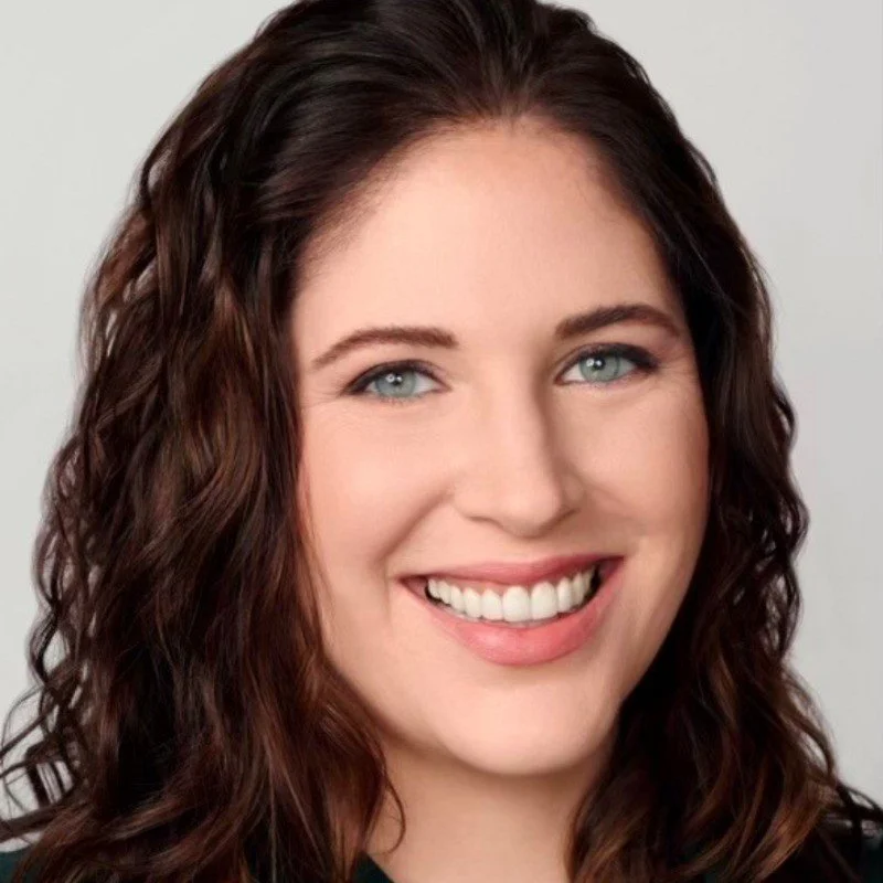 A close-up portrait of a young woman with long, curly brown hair and blue eyes, smiling at the camera.