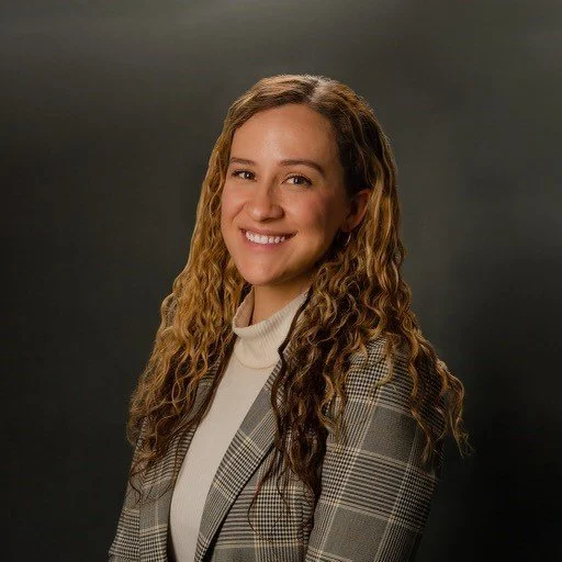 A young woman with long, curly hair smiling at the camera, wearing a plaid blazer over a turtleneck, against a dark gray background.