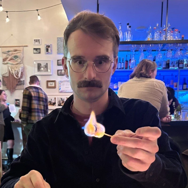 A man with glasses and a mustache holding a lit marshmallow on a stick in a bar or restaurant with people in the background.