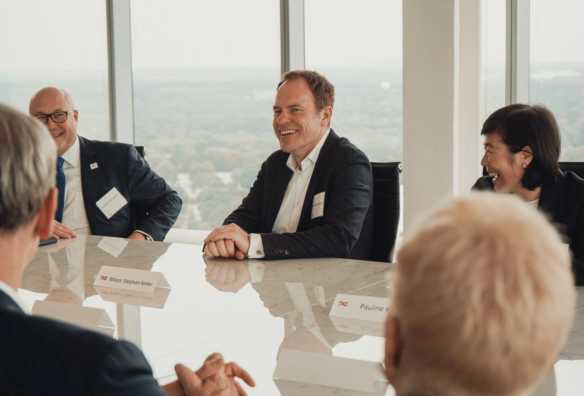 Group of people in business attire sitting around a conference table, smiling and engaging in conversation, with large windows and cityscape in the background.
