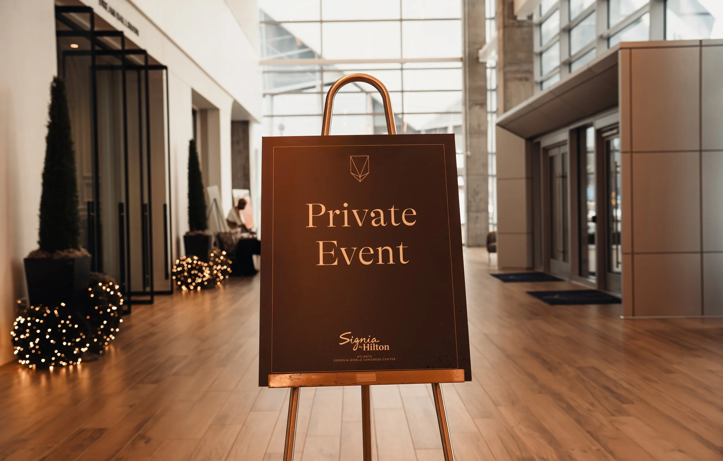 A sign reading 'Private Event' on an easel inside a modern building with tall glass windows, hardwood floors, and potted plants.