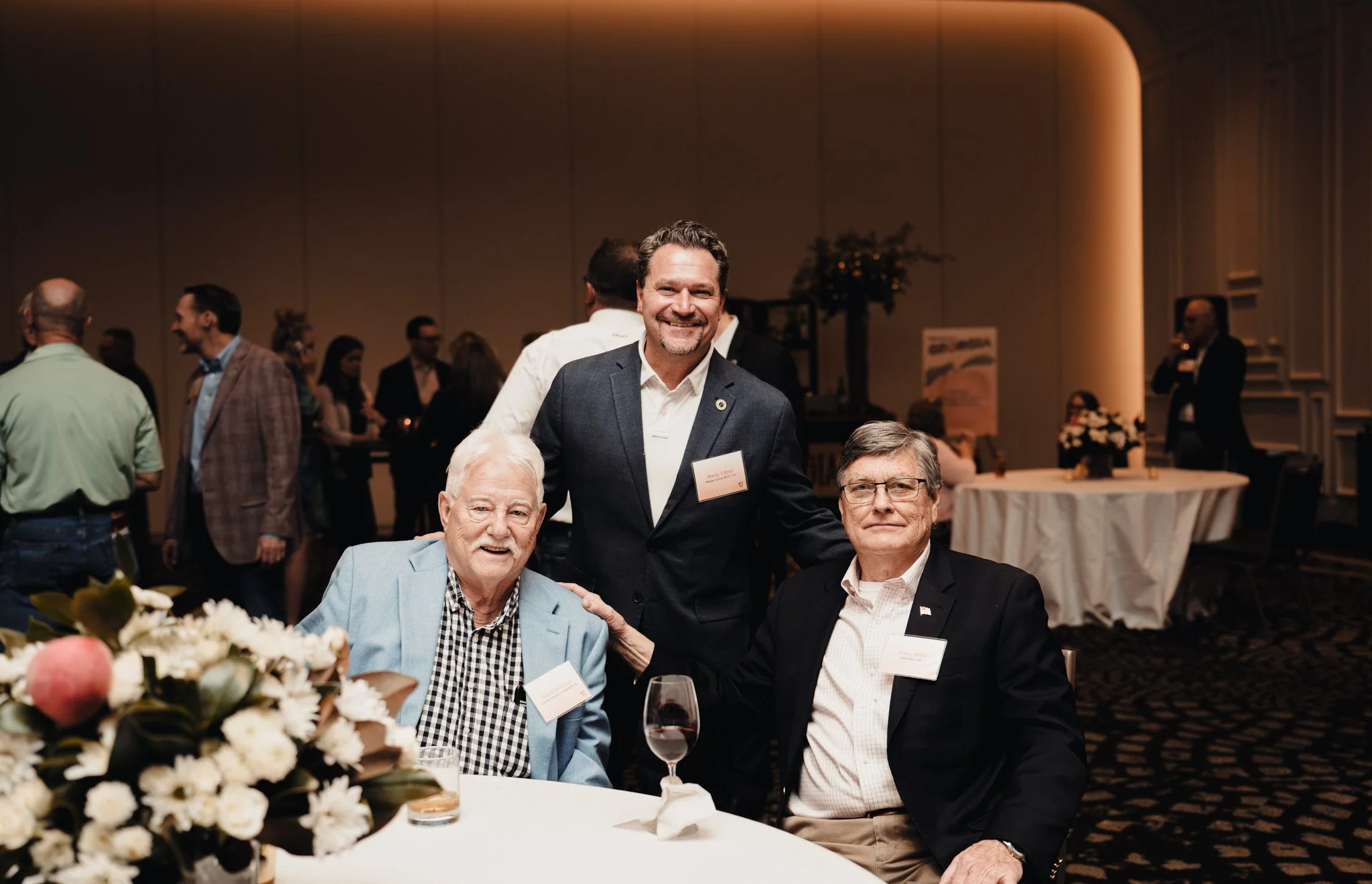 A group of men at a formal event, sitting at a table with a floral centerpiece, with a man in a dark suit standing behind two seated men, all smiling for the photo.