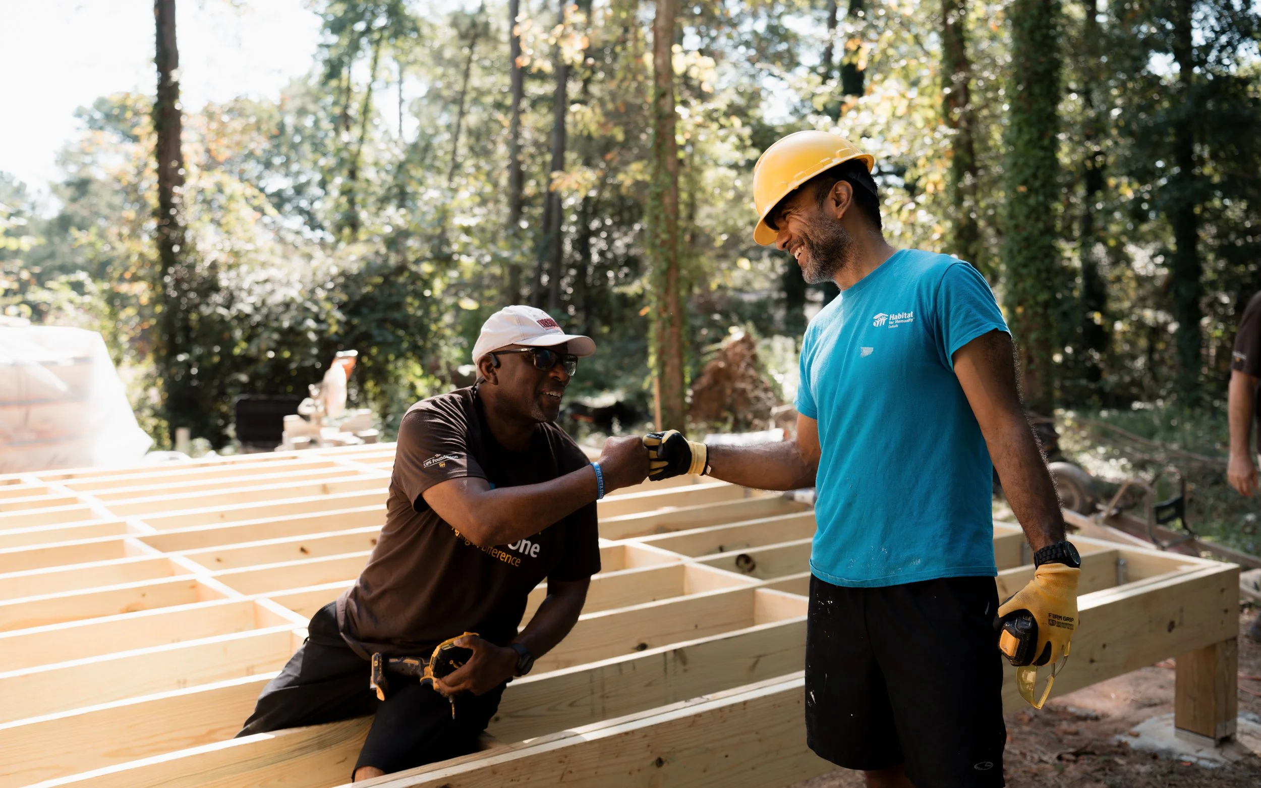 Two construction workers shaking hands on a wooden frame of a building in a wooded outdoor area. One man is kneeling, wearing sunglasses, a white cap, and a black shirt. The other man is standing, wearing a yellow hard hat, a blue shirt, and holding a power drill.
