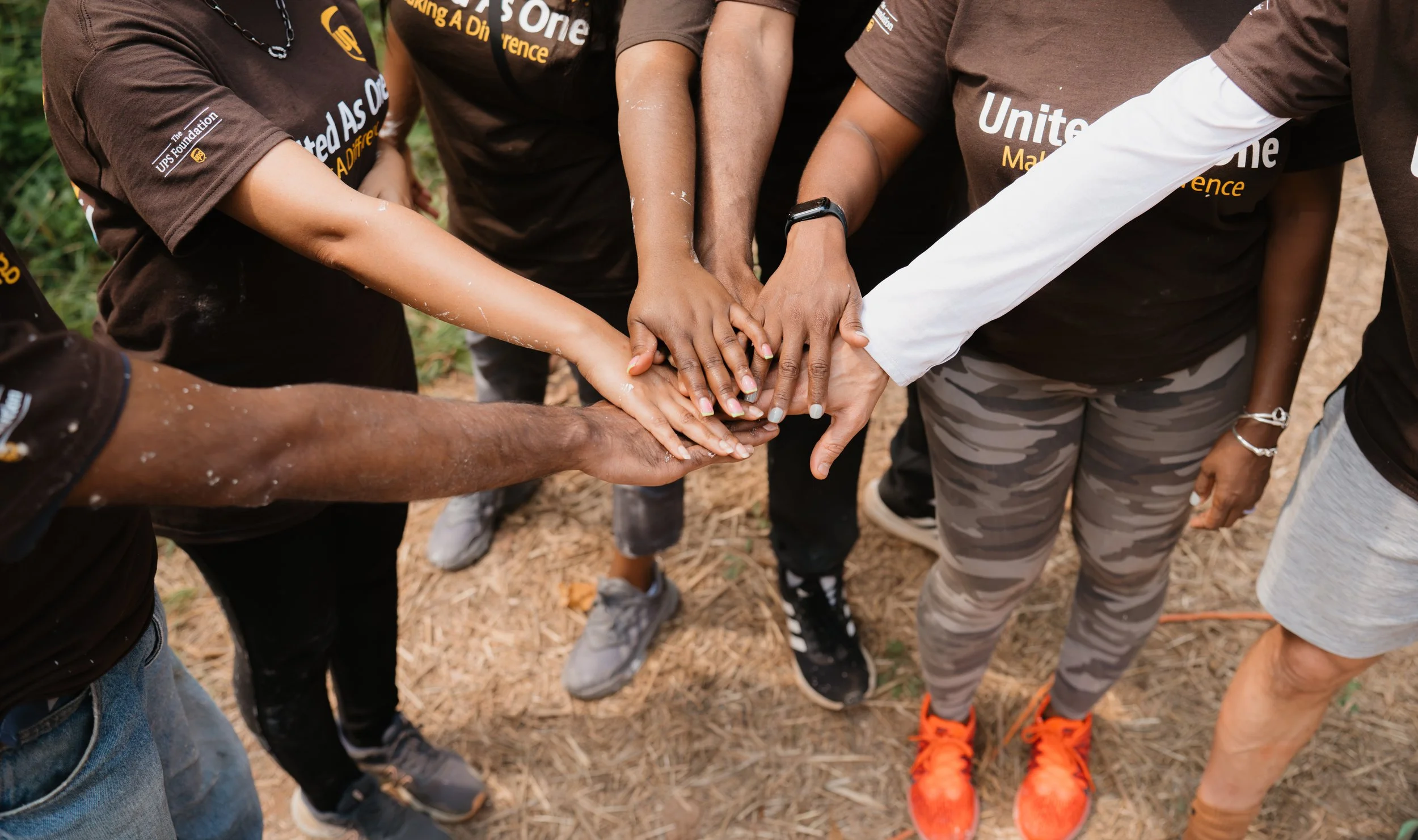 Multiple people with varying skin tones from different backgrounds place their hands together in the center in a gesture of teamwork outdoors on dirt ground.