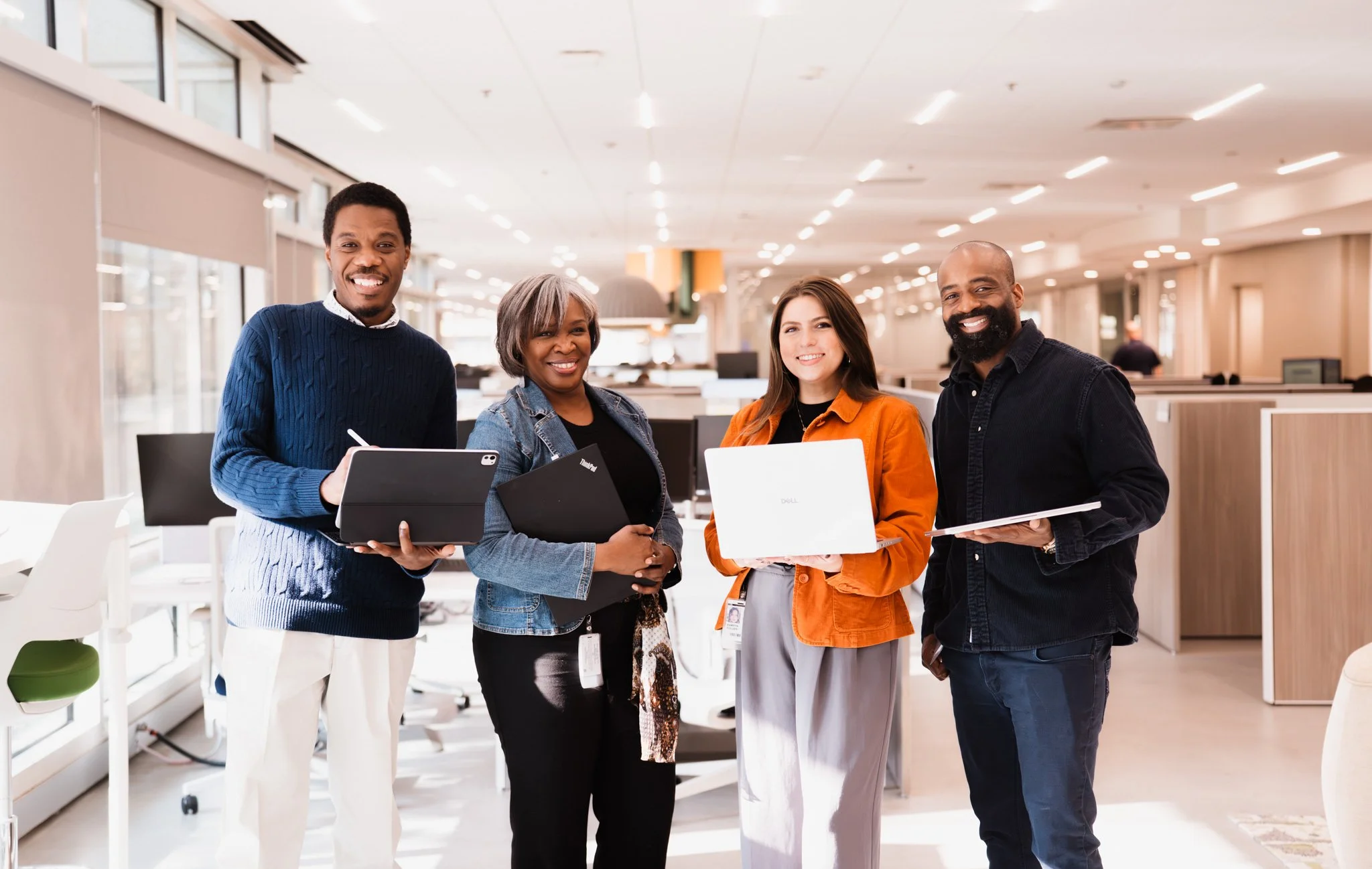 Four diverse professionals standing together in an office, smiling and holding laptops and tablets.