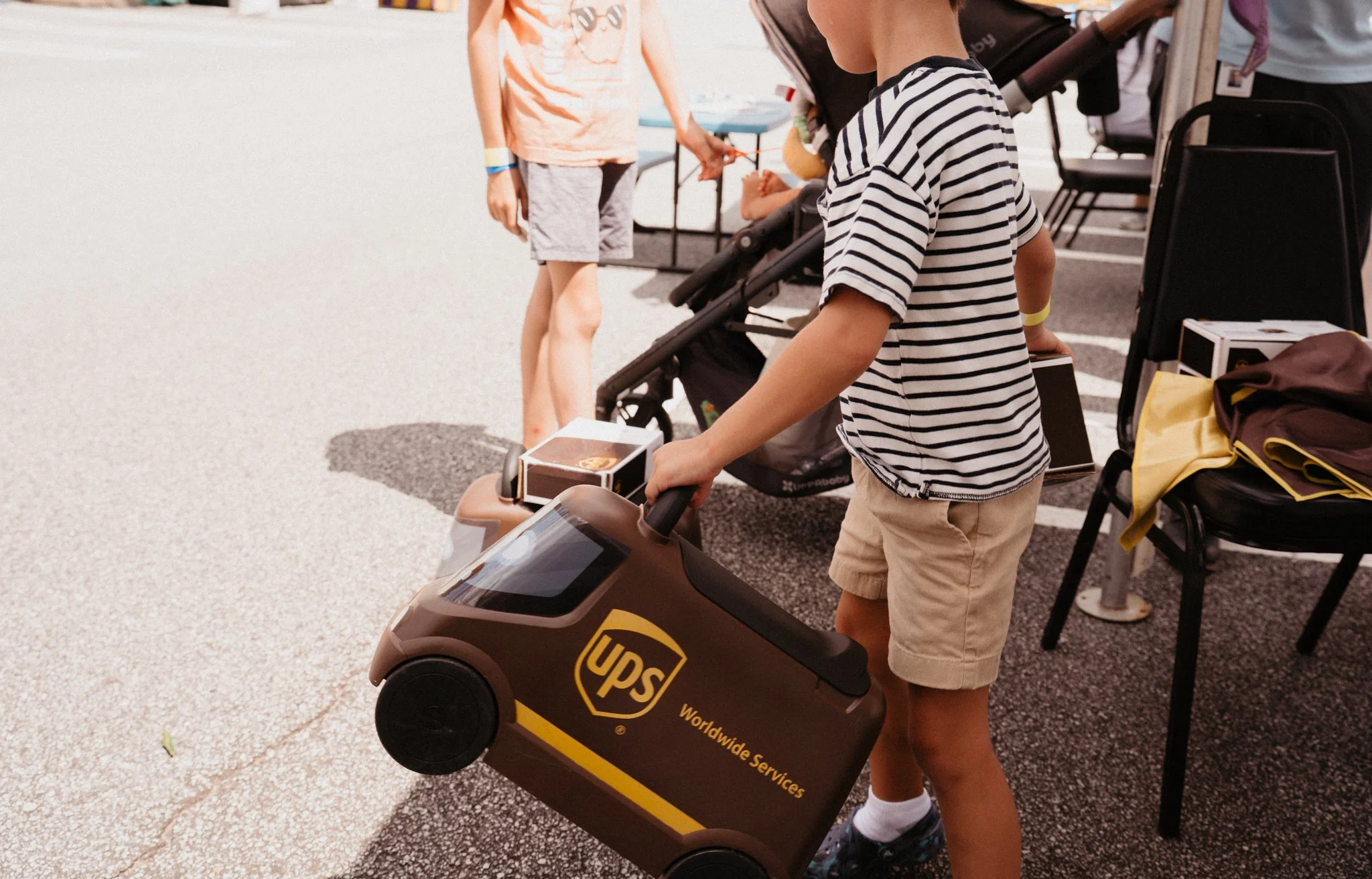 Child in striped shirt and beige shorts riding a toy motorcycle with a UPS logo, others in the background near a stroller and outdoor tables.