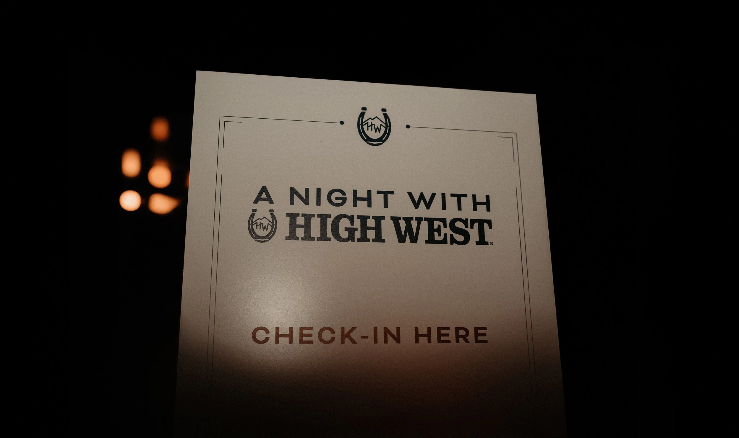 A sign with the logo of 'A Night with High West', indicating a check-in area in a dark environment with some warm light sources in the background.