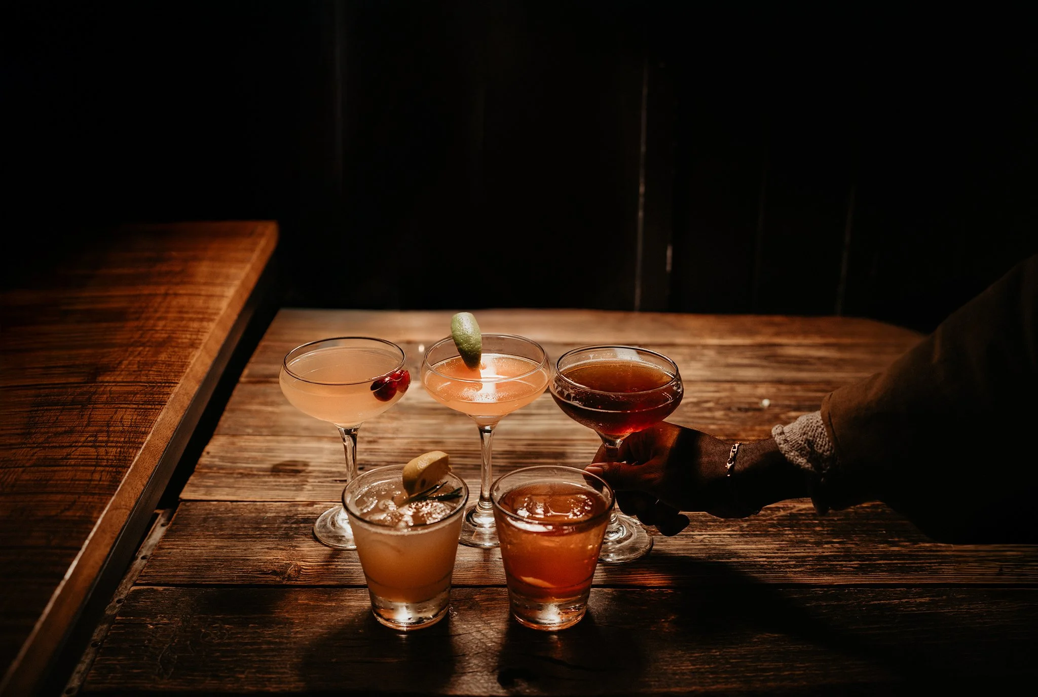 Four cocktails on a wooden bar, with one lime wedge and a cherry garnish, and a hand reaching for a glass on the right.