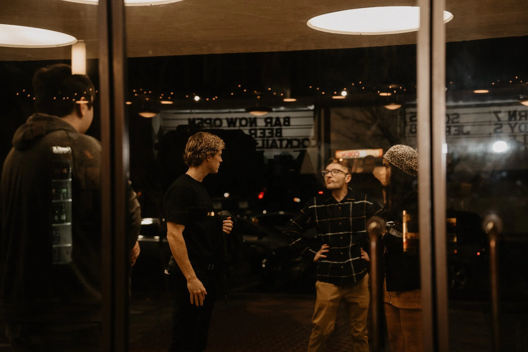 Group of four people inside a restaurant or cafe, engaged in conversation, viewed through a glass window at night.