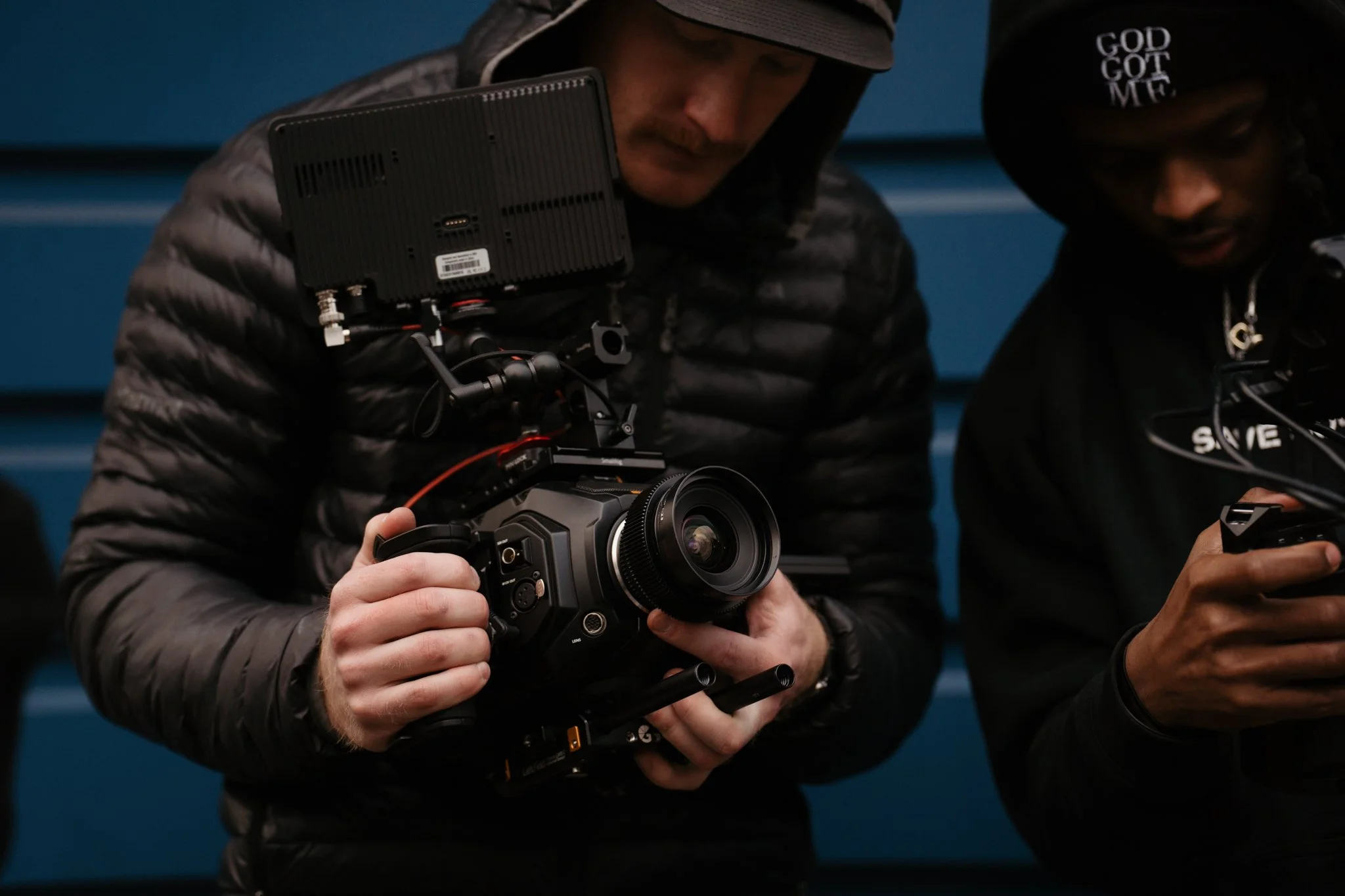 Two individuals look at a camera and a smartphone, with one holding professional video equipment and the other viewing a device. They are dressed in dark clothing with hoodies.