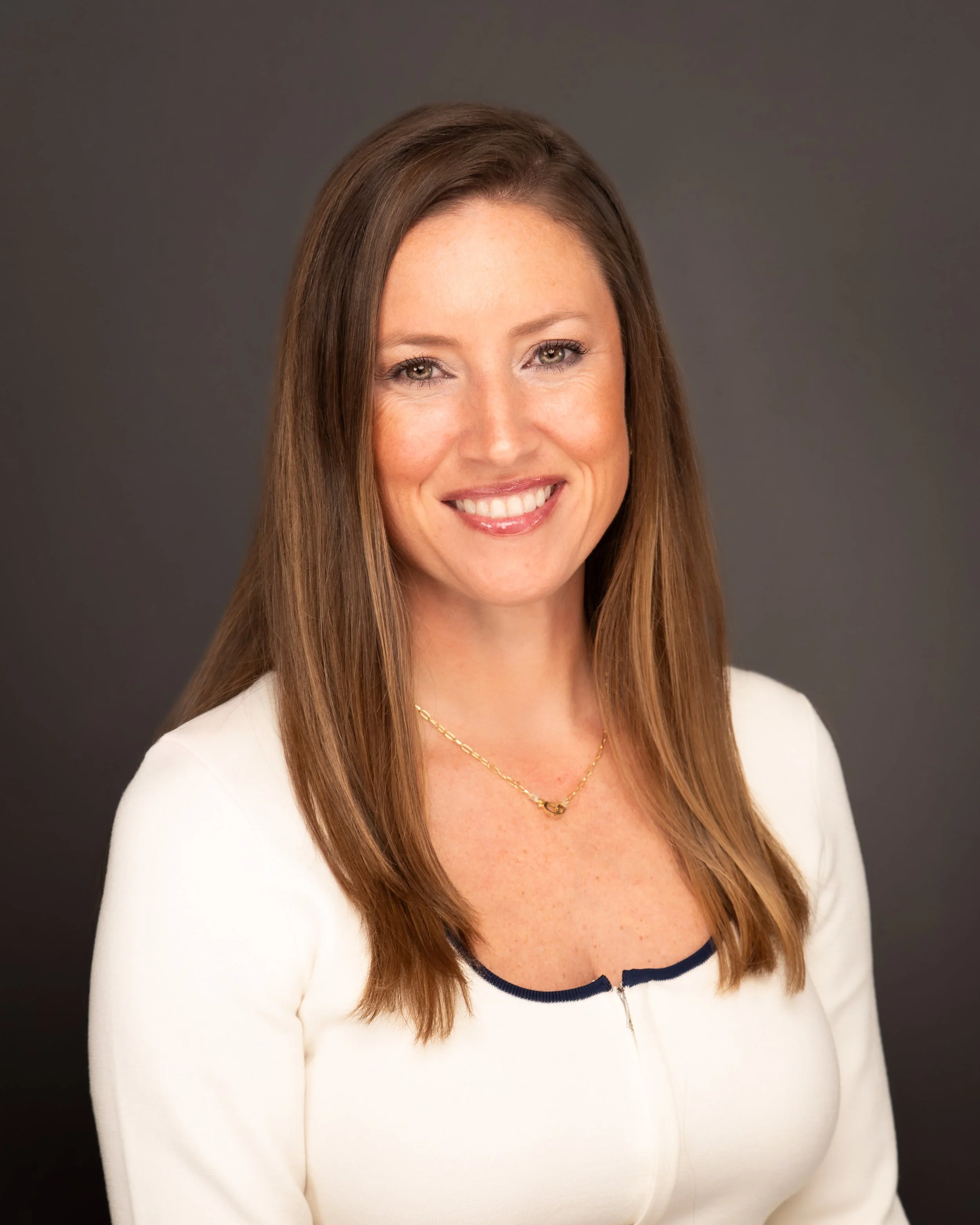 A woman with brown hair, wearing a white sweater with navy trim, gold jewelry, and a friendly smile, posed against a gray background.