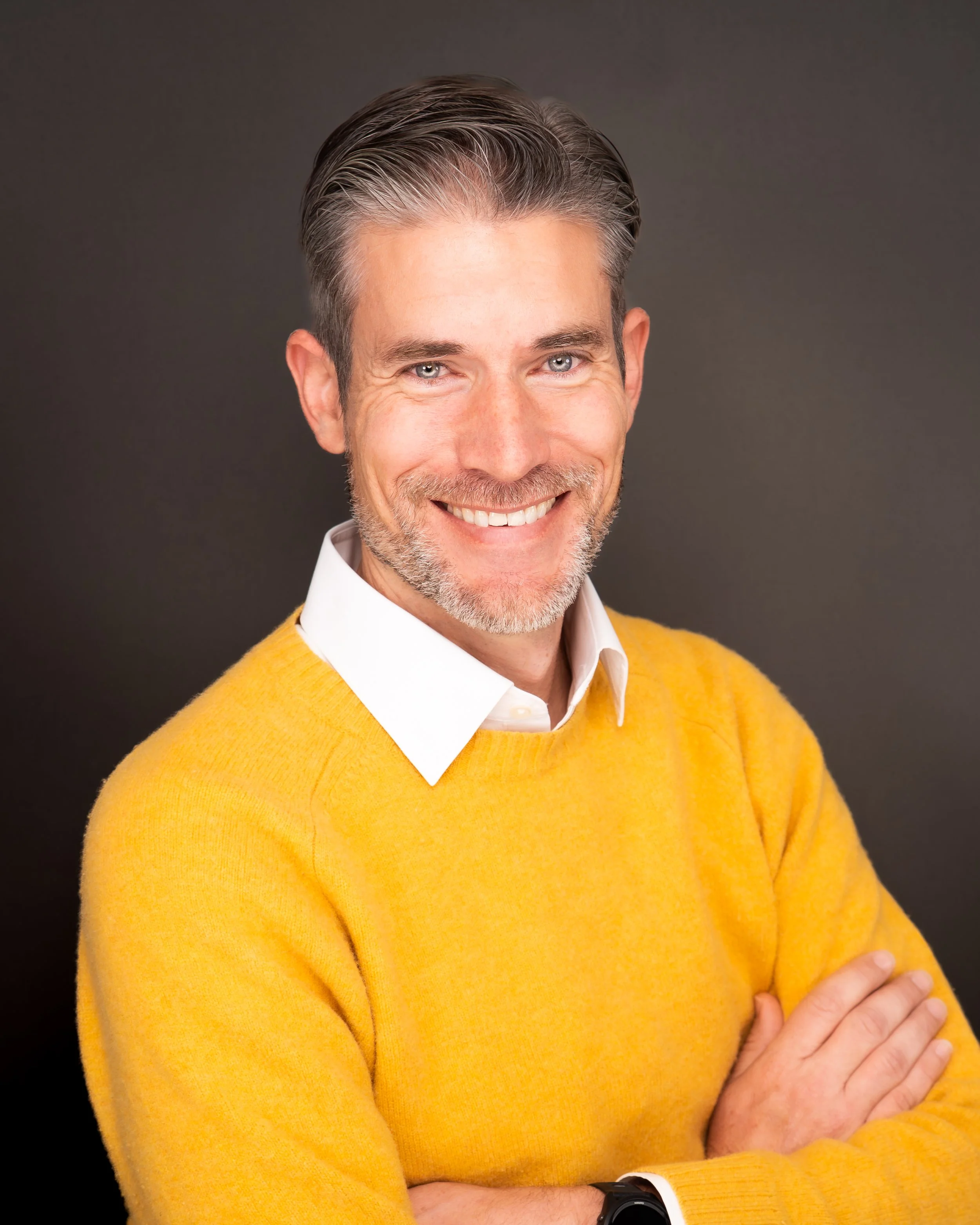 A smiling middle-aged man with gray hair and a beard, wearing a white shirt and a yellow sweater, stands against a dark background with crossed arms.