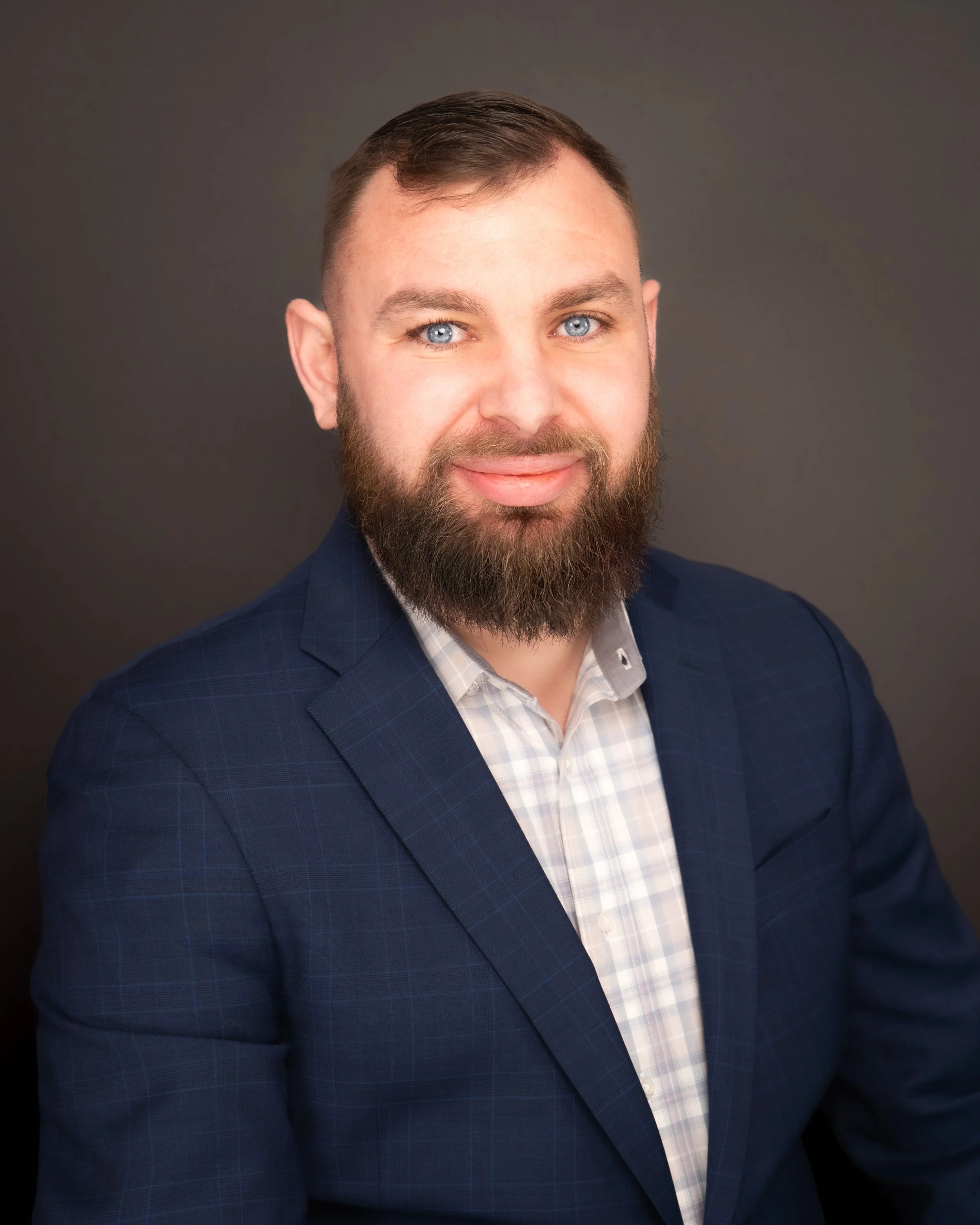 A man with a beard and blue eyes wearing a dark blue checkered suit jacket and a light-colored dress shirt, smiling against a plain dark background.