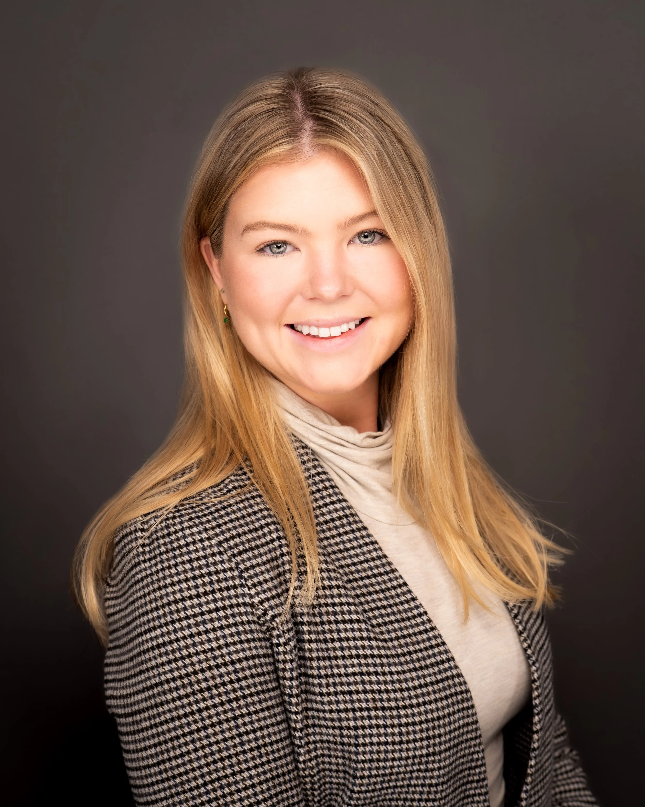 A portrait of a young woman with long blonde hair, blue eyes, and a friendly smile, wearing a checkered blazer and a beige scarf, against a dark background.