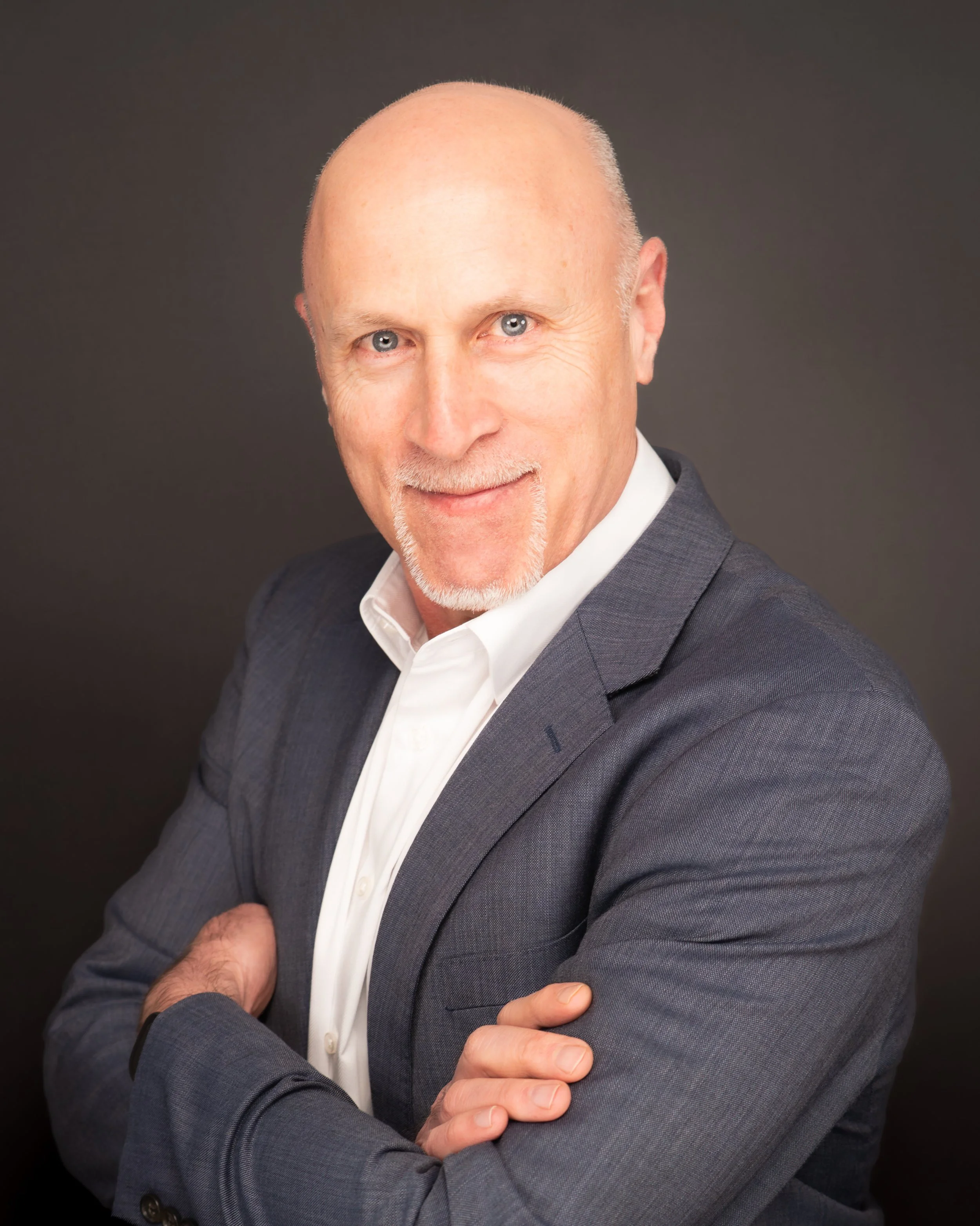 A bald man with blue eyes and a goatee wearing a dark gray suit jacket and a white dress shirt, posing with folded arms against a dark background.