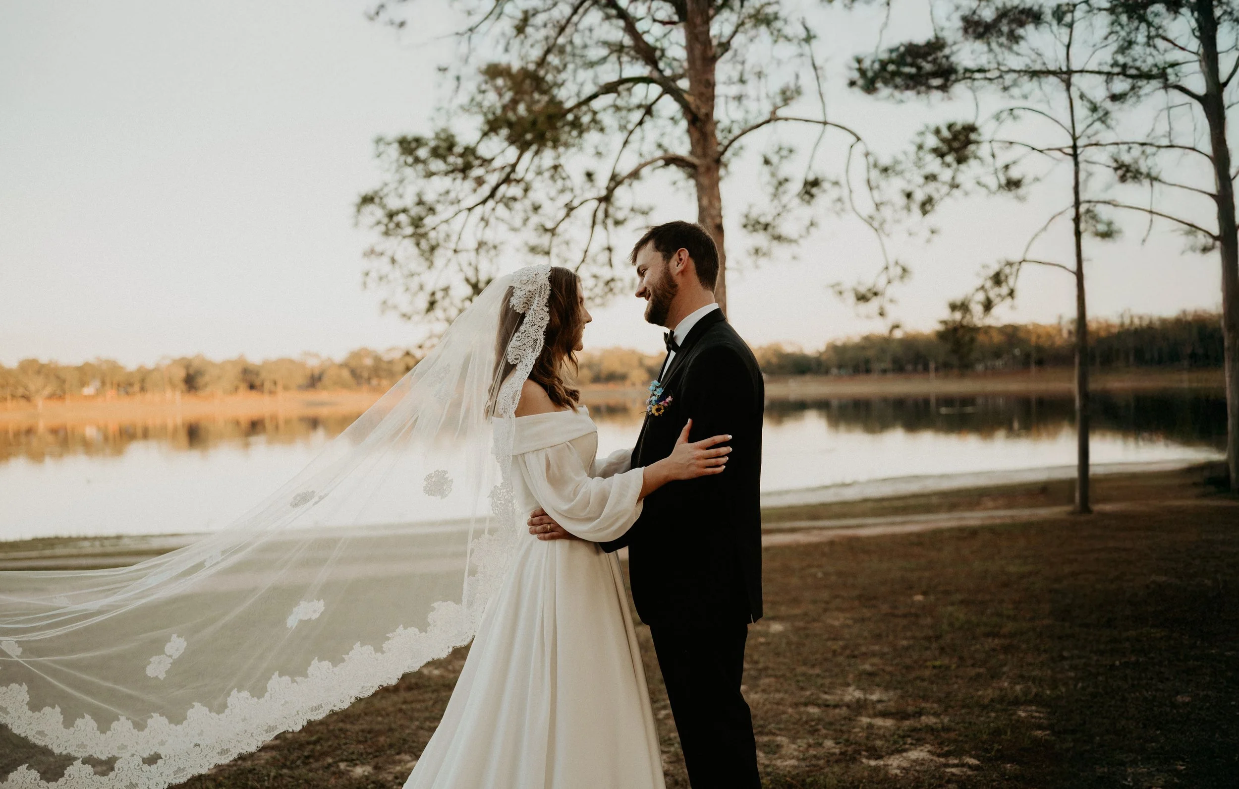 A bride and groom standing face-to-face by a lake, smiling at each other during sunset, with trees and a calm water background.