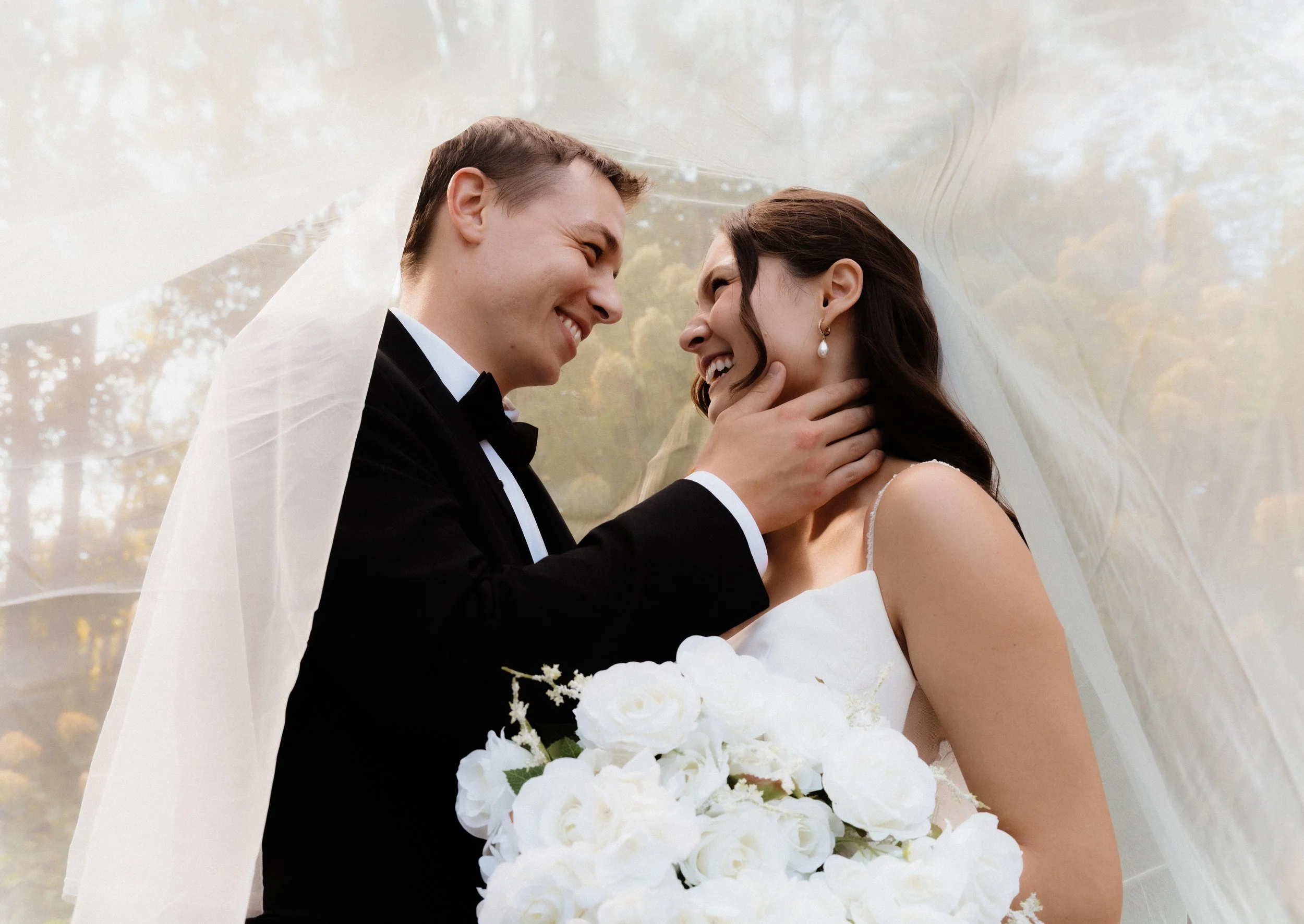 A bride and groom smiling and touching noses in an outdoor wedding setting, with the bride holding a bouquet of white flowers and wearing earrings, and the groom in a tuxedo.