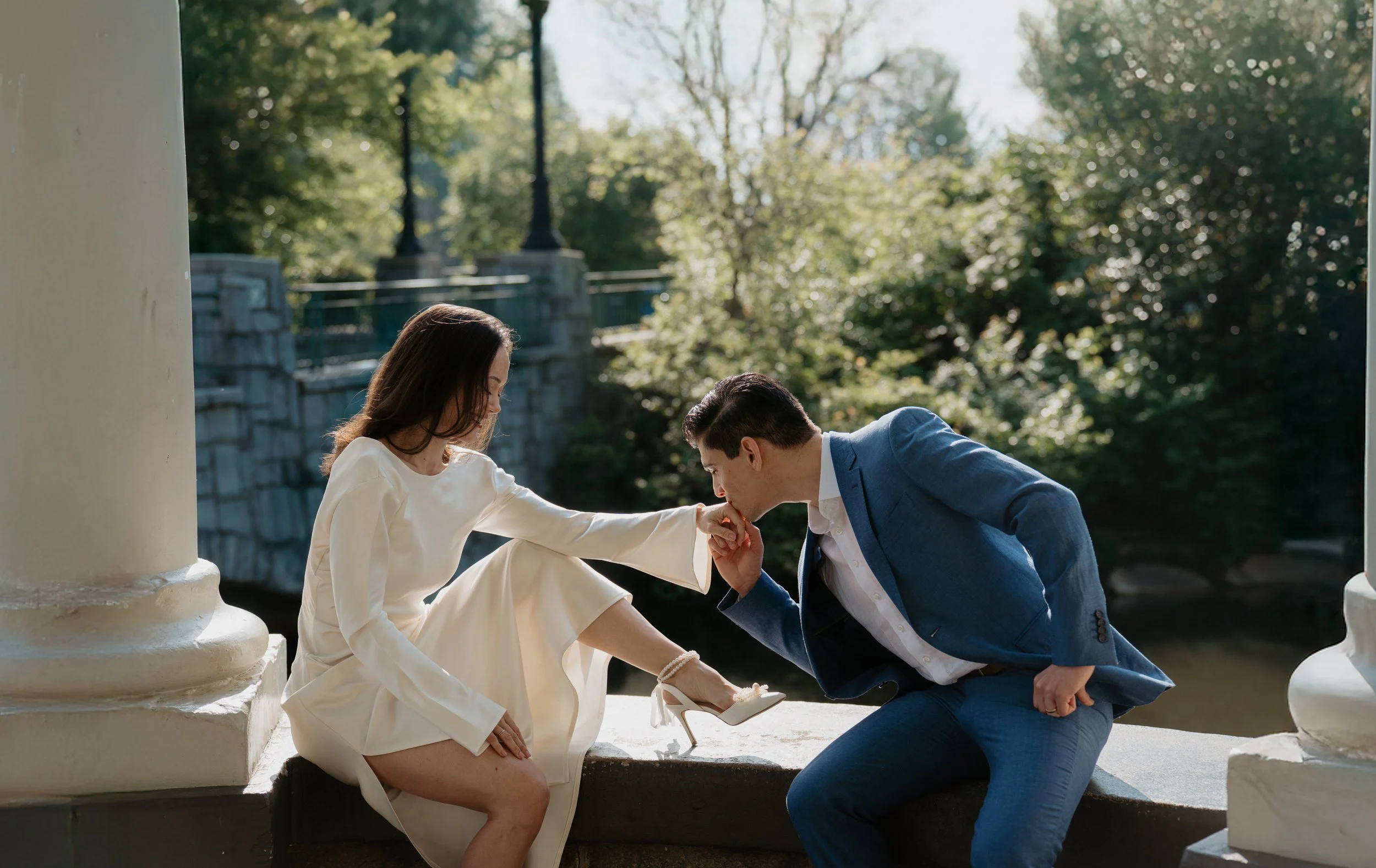 A man kneeling and kissing the hand of a woman dressed in a white dress, sitting on a stone ledge with an outdoor park background.