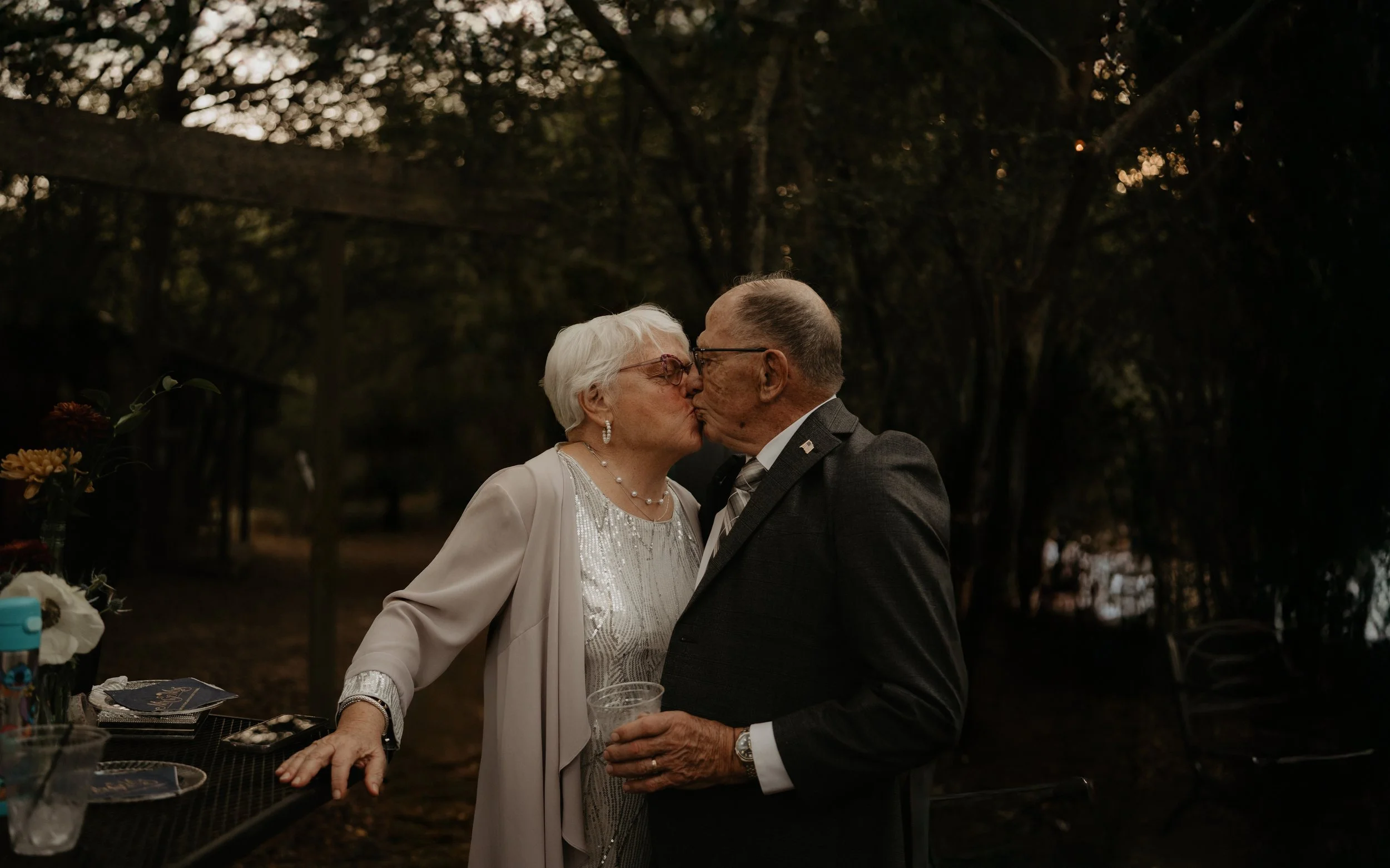 An elderly couple sharing a kiss at an outdoor event during sunset, with trees in the background. The woman is wearing a white outfit and pearl jewelry, and the man is in a dark suit with glasses.