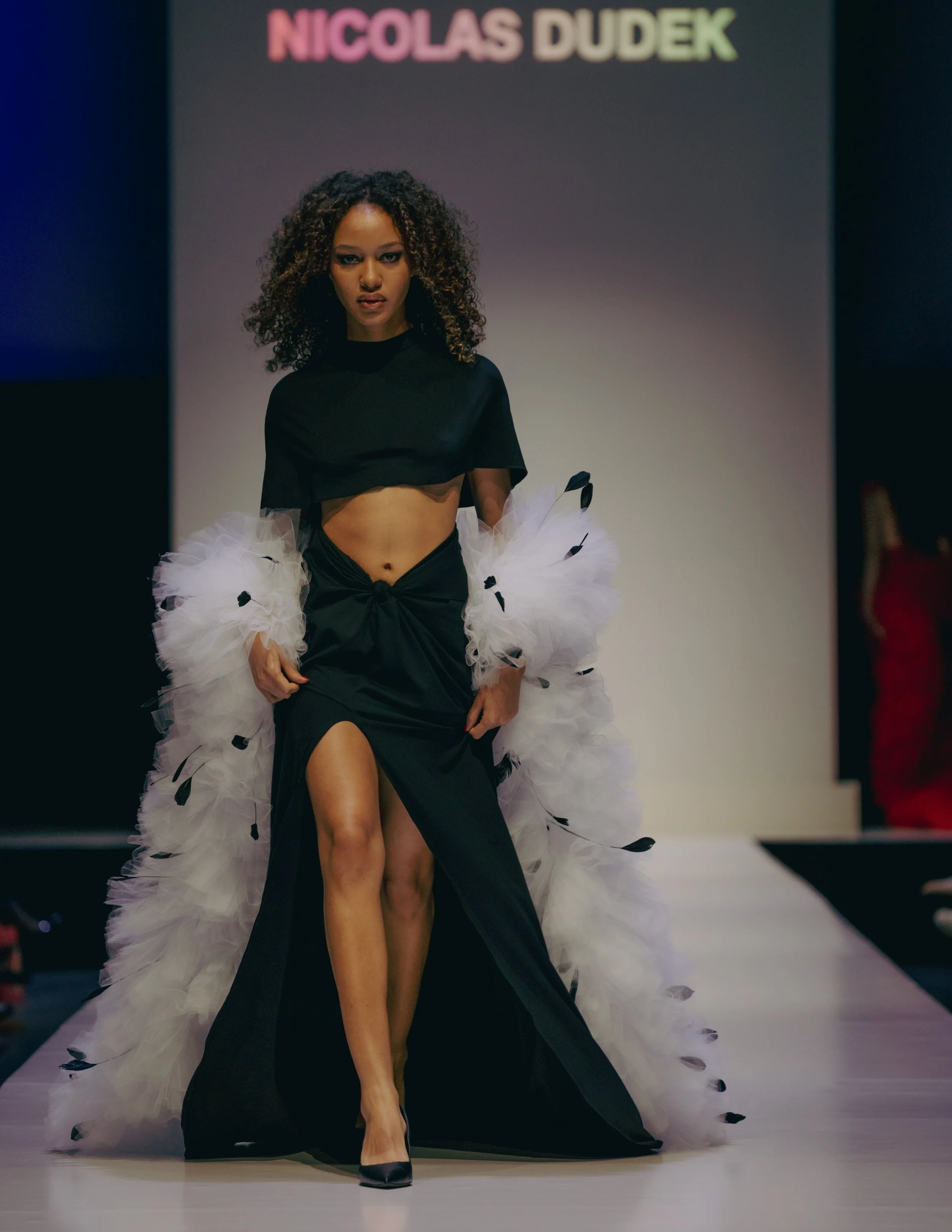 A female model walking runway wearing a black dress with a thigh-high slit, paired with black heels, and fluffy white and black feathered accessories. Behind her, a signature 'Nicolas Dudek' is displayed on a gray background.