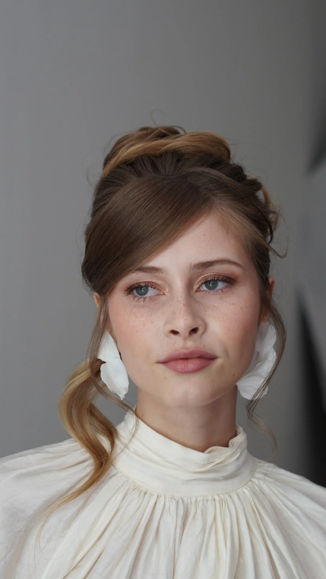 A woman with styled hair, blue eyes, and freckles on her face, wearing a white ruffled shirt and white earrings, posing against a plain background.