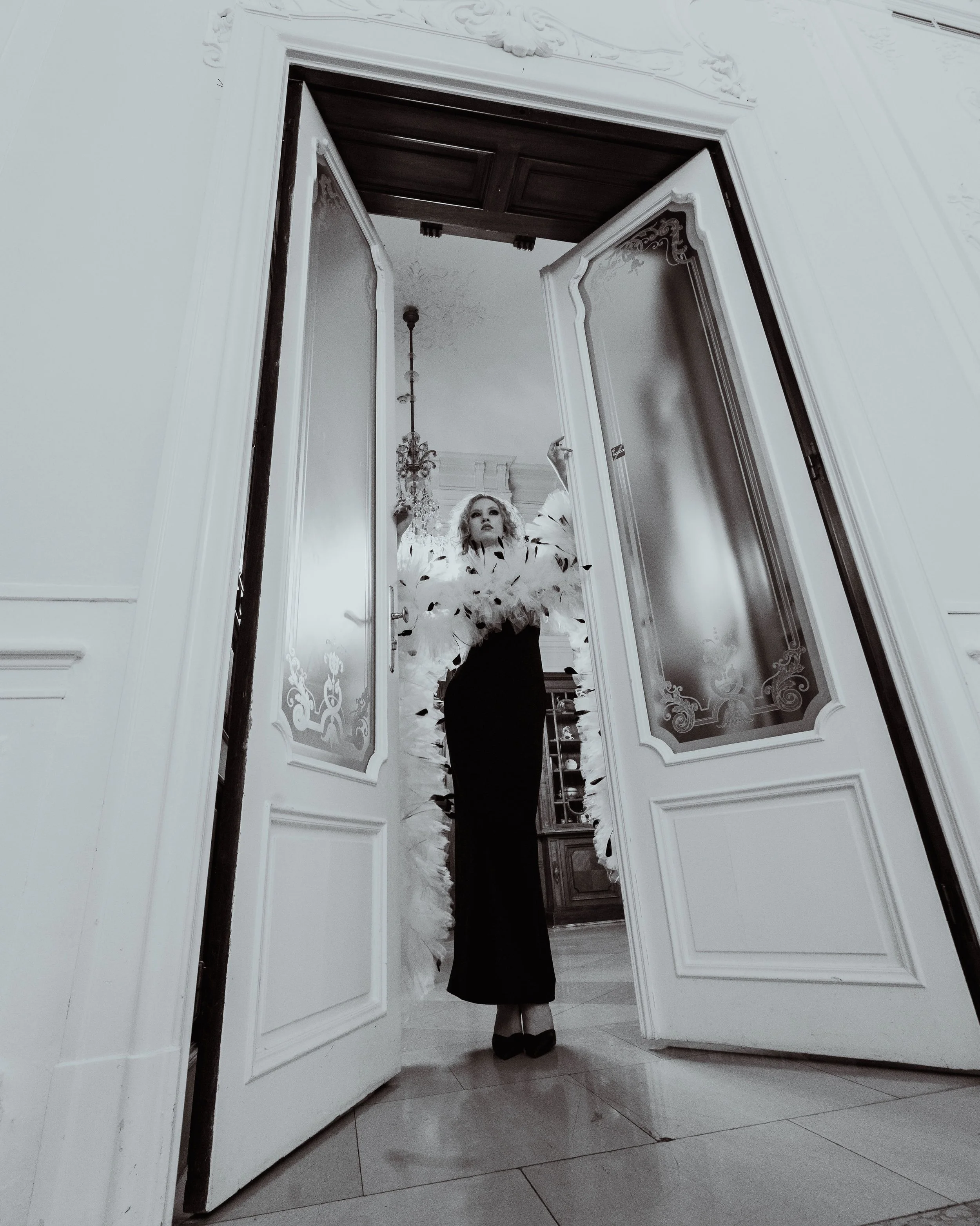 A woman standing in a hallway is seen through an open door mirror, dressed in formal attire with a puffed sleeve dress and high heels. The hallway has elegant decor, including a chandelier and wall moldings.