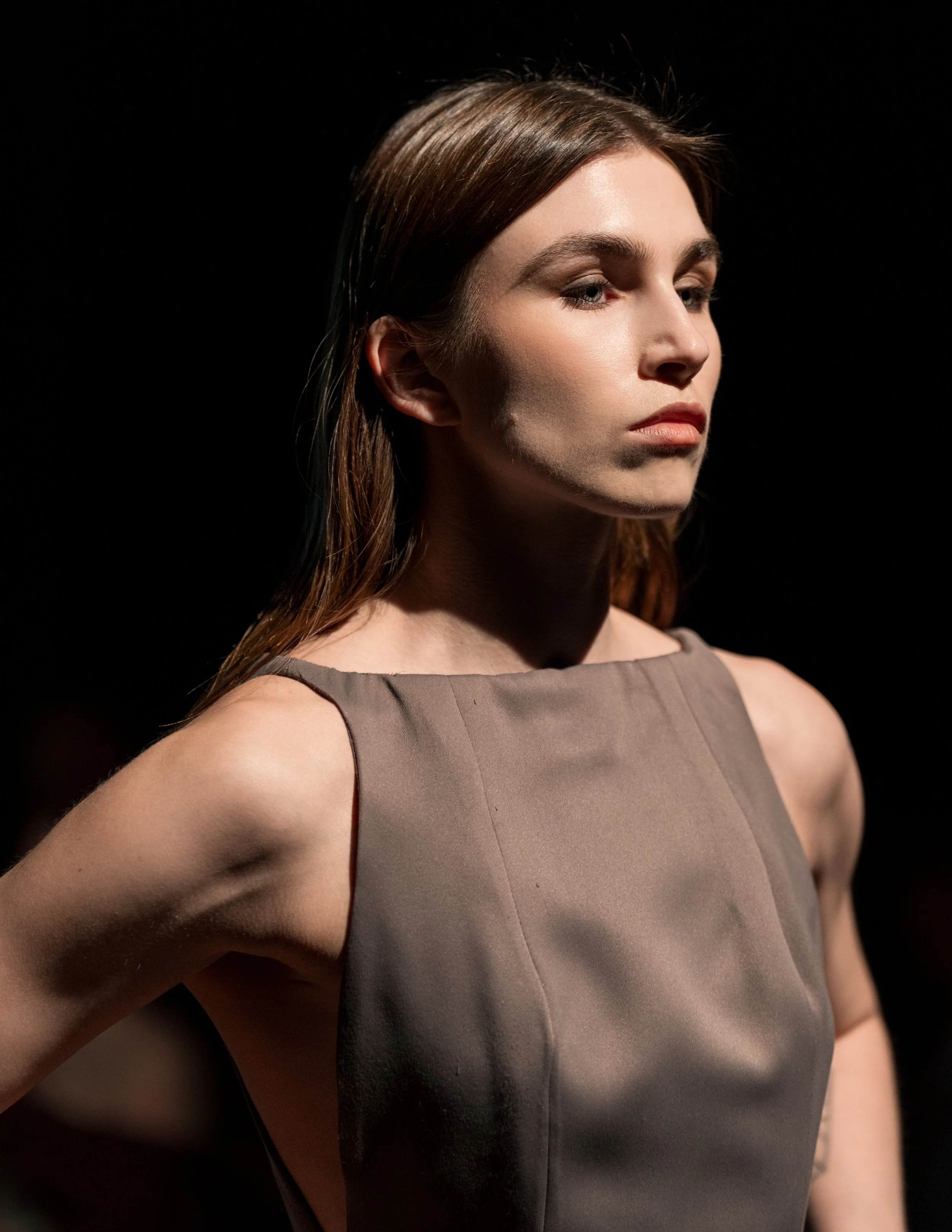 Close-up of a woman with brown hair, wearing a sleeveless taupe dress, looking to her left, against a black background.