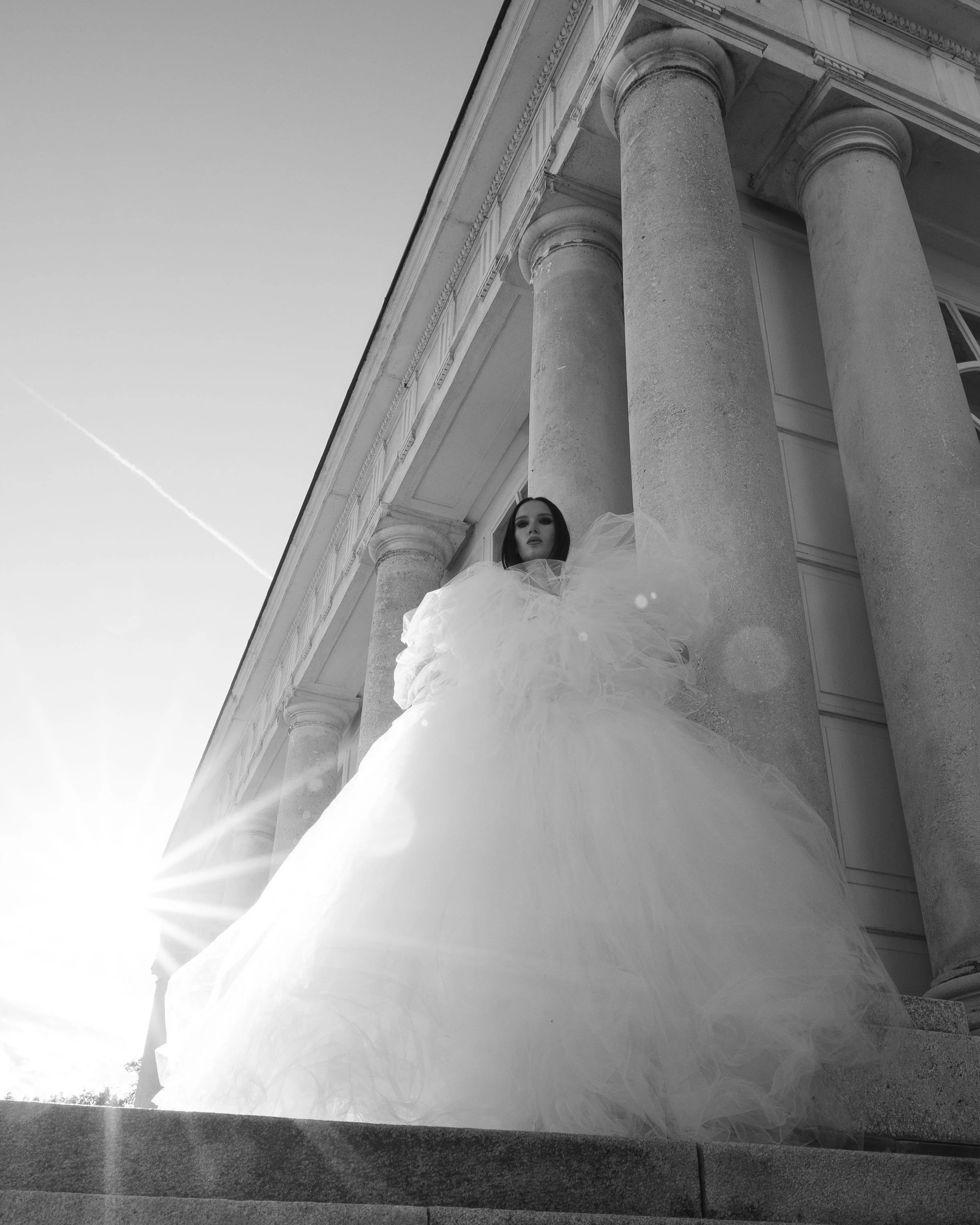 Black and white photo of a woman with dark hair in a white dress standing behind tall stone columns of a classical building, looking at the camera.
