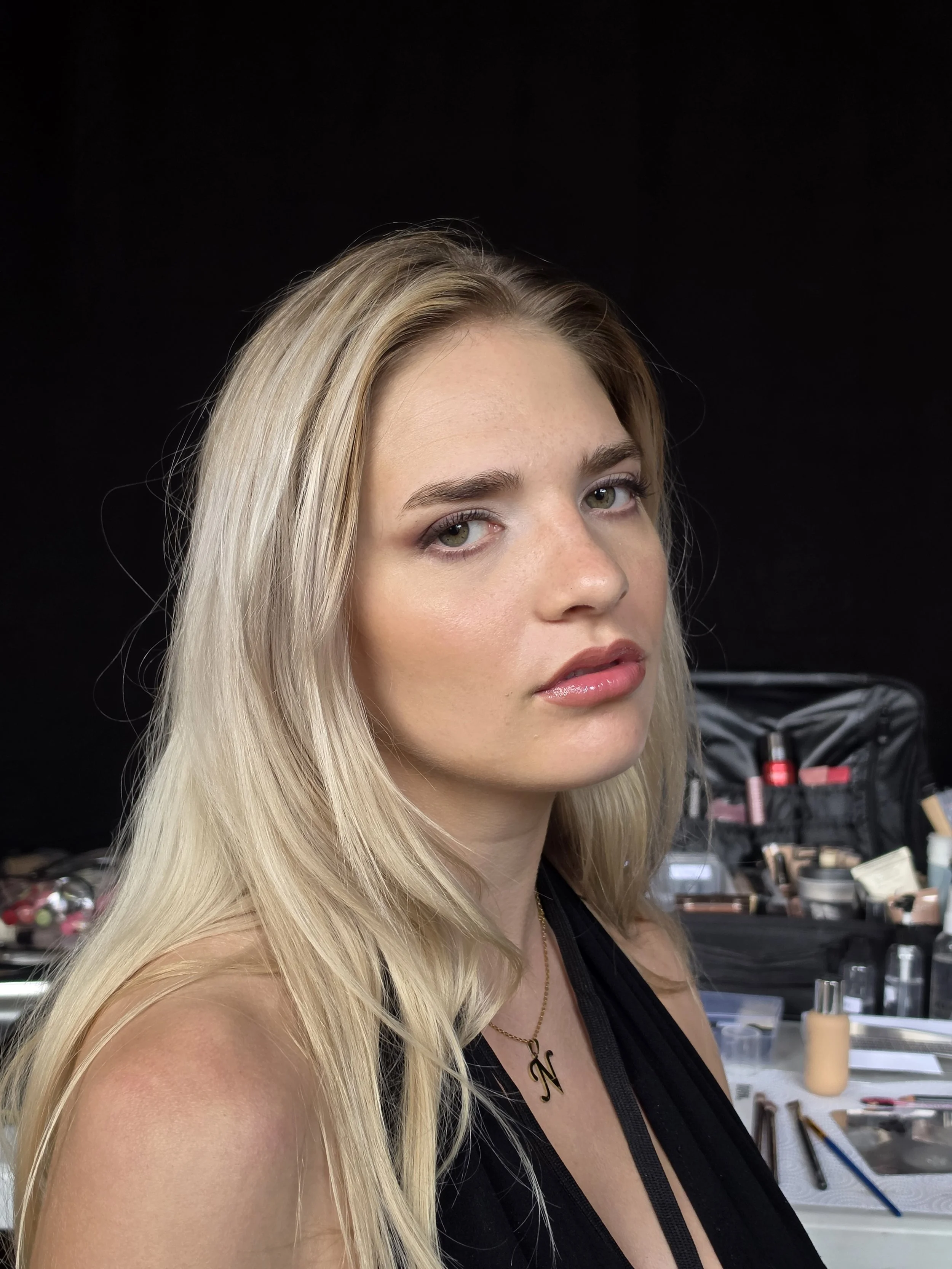 A woman with long blonde hair and green eyes at a makeup station, surrounded by makeup products and tools.