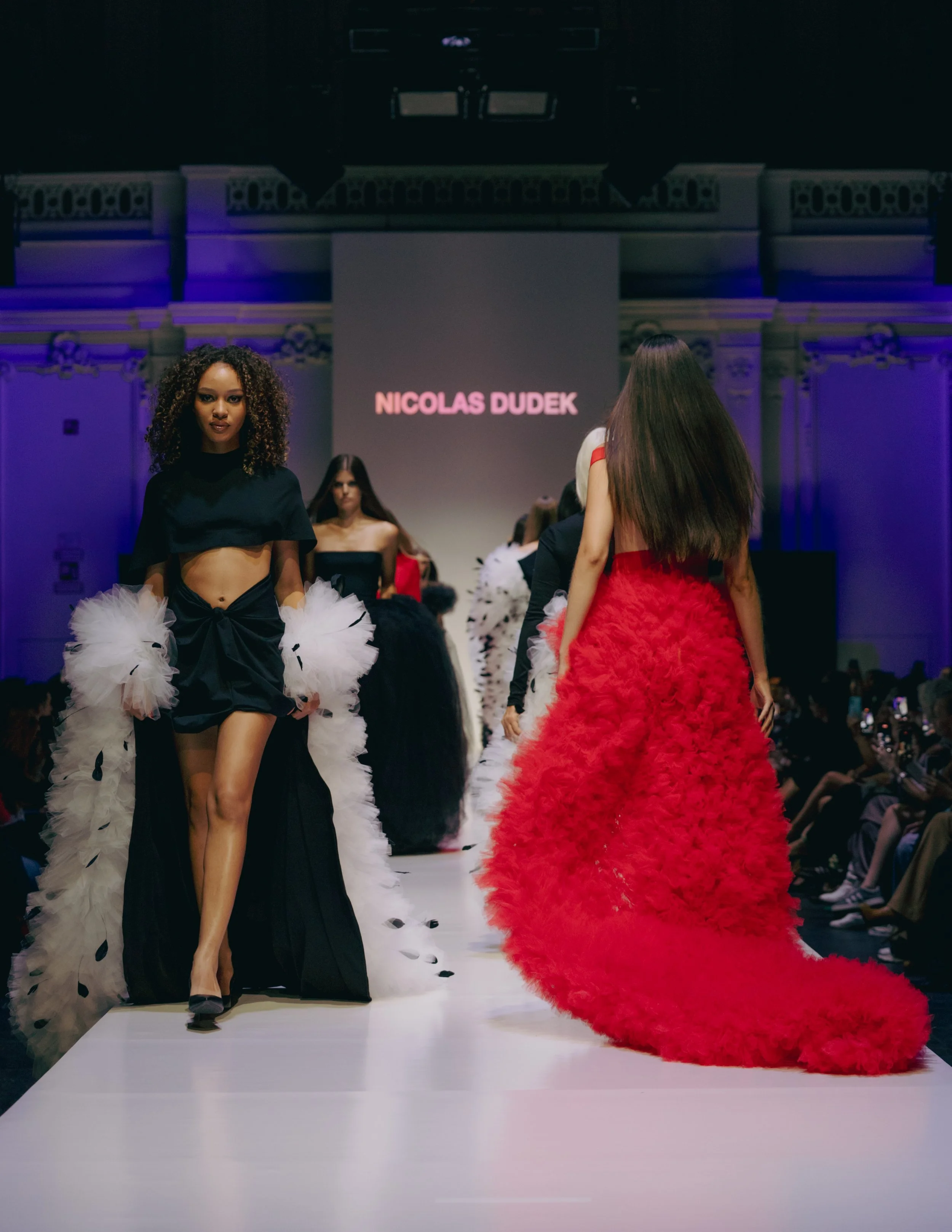 Models walking on the runway at a fashion show, with a large screen behind displaying the name Nicolas Dudek. The models are wearing high-fashion outfits, some with feathered designs.